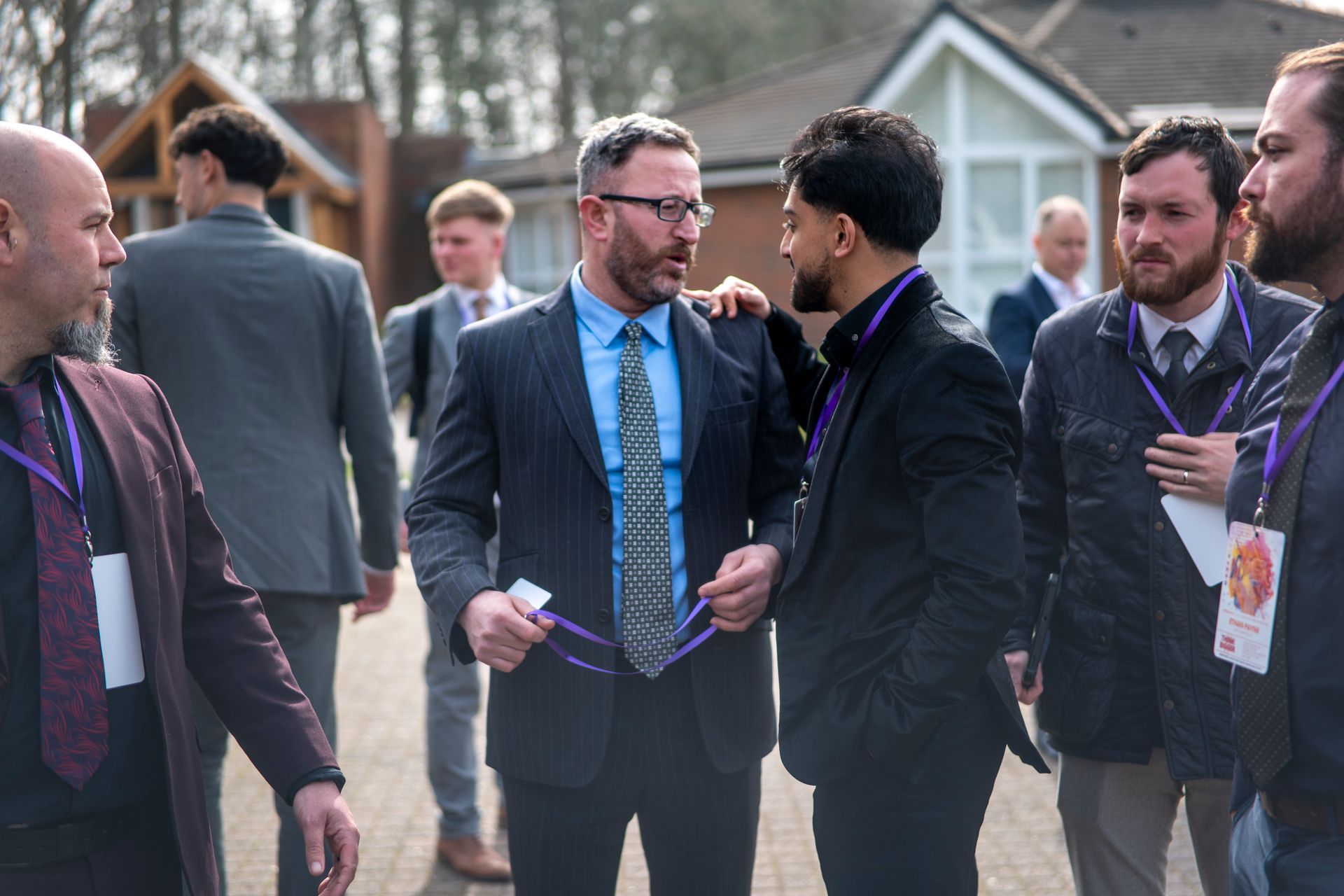 A group of individuals in professional attire, some wearing event lanyards, interacting outdoors at a venue entrance.