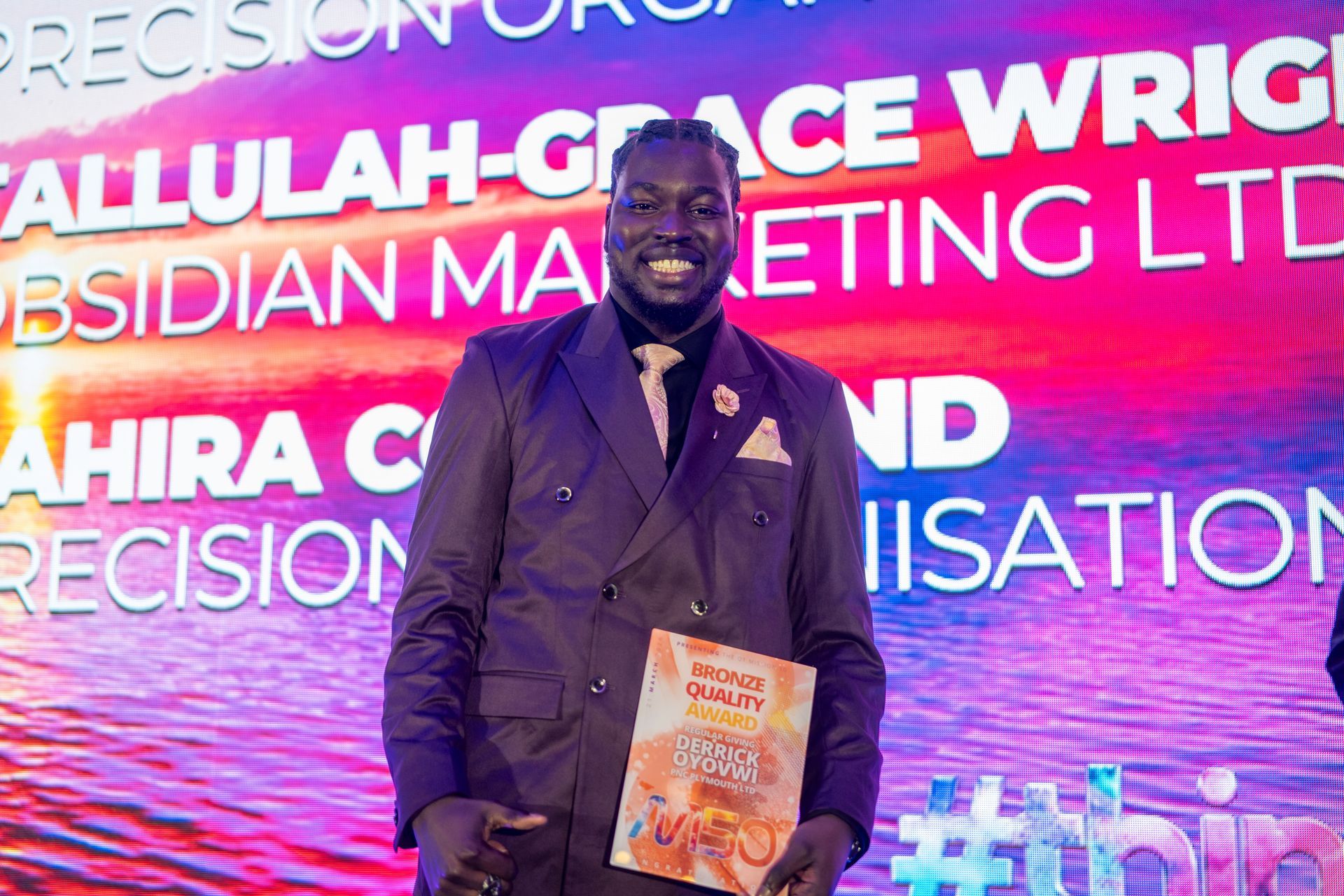 A smiling individual stands on a stage holding an award against a vibrant, sunset-colored backdrop with text.