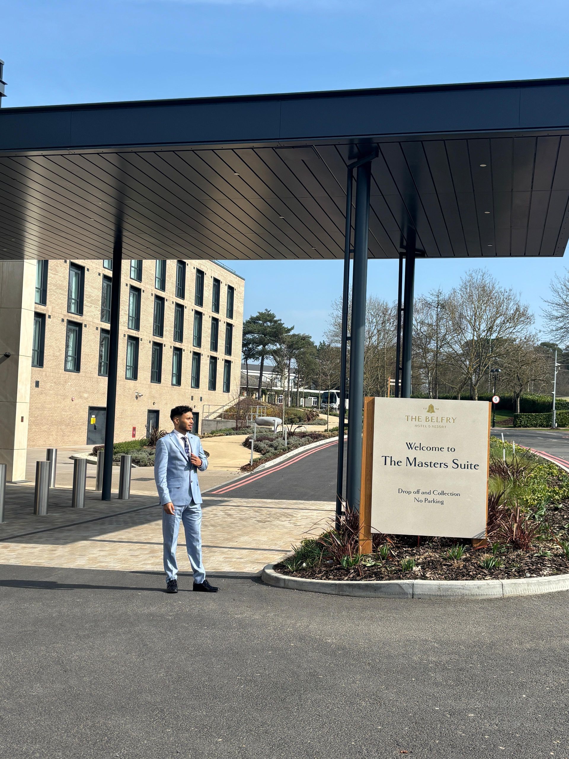 A person in a light blue suit stands under a modern, patterned metal awning in front of a tan building and signage.