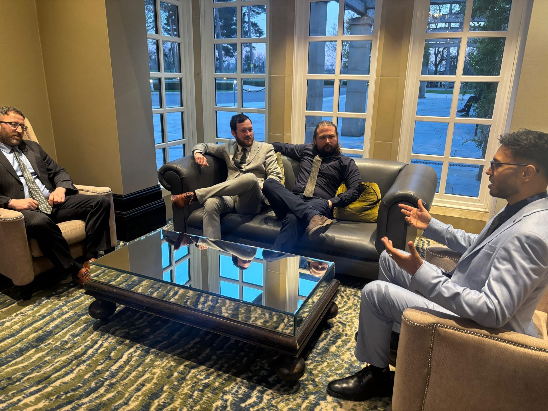 Four people in business casual attire sit in a lounge with floor-to-ceiling windows, engaged in a group conversation.