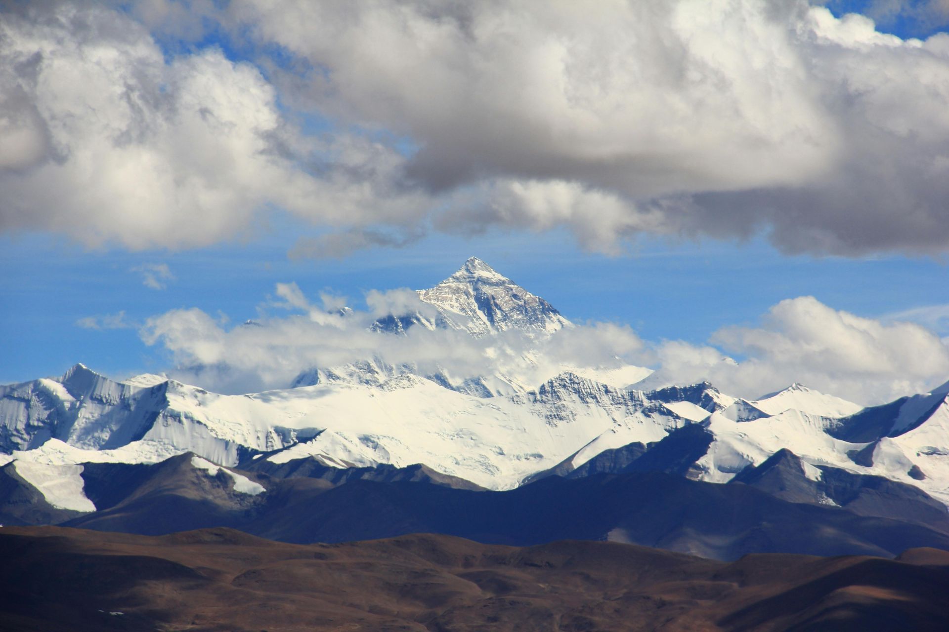 Snow-capped Mount Everest rises majestically above a mountain range, partially framed by soft clouds in a blue sky.