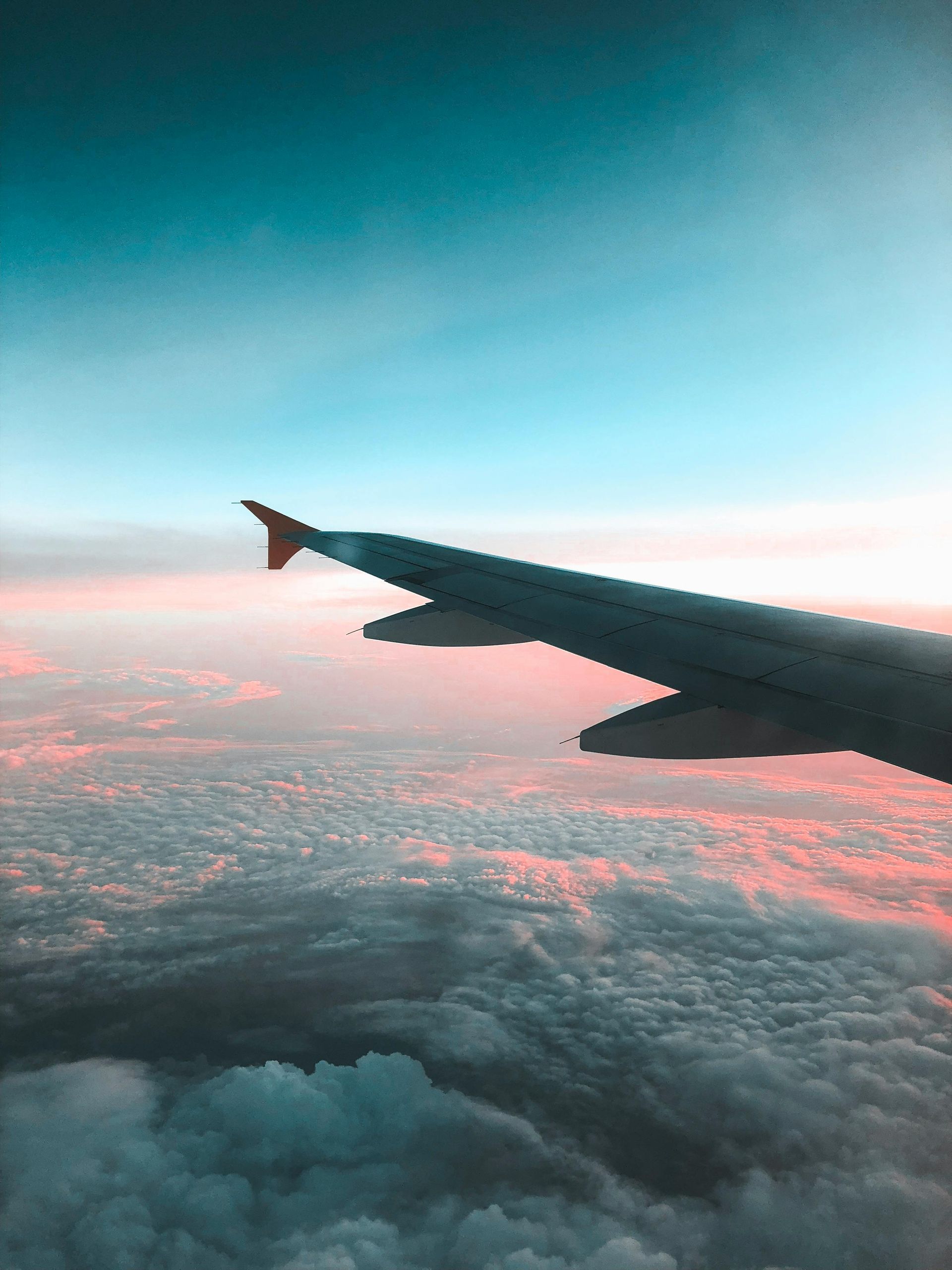 View from an airplane window showing the wing above a blanket of clouds during a pink and blue sunset.