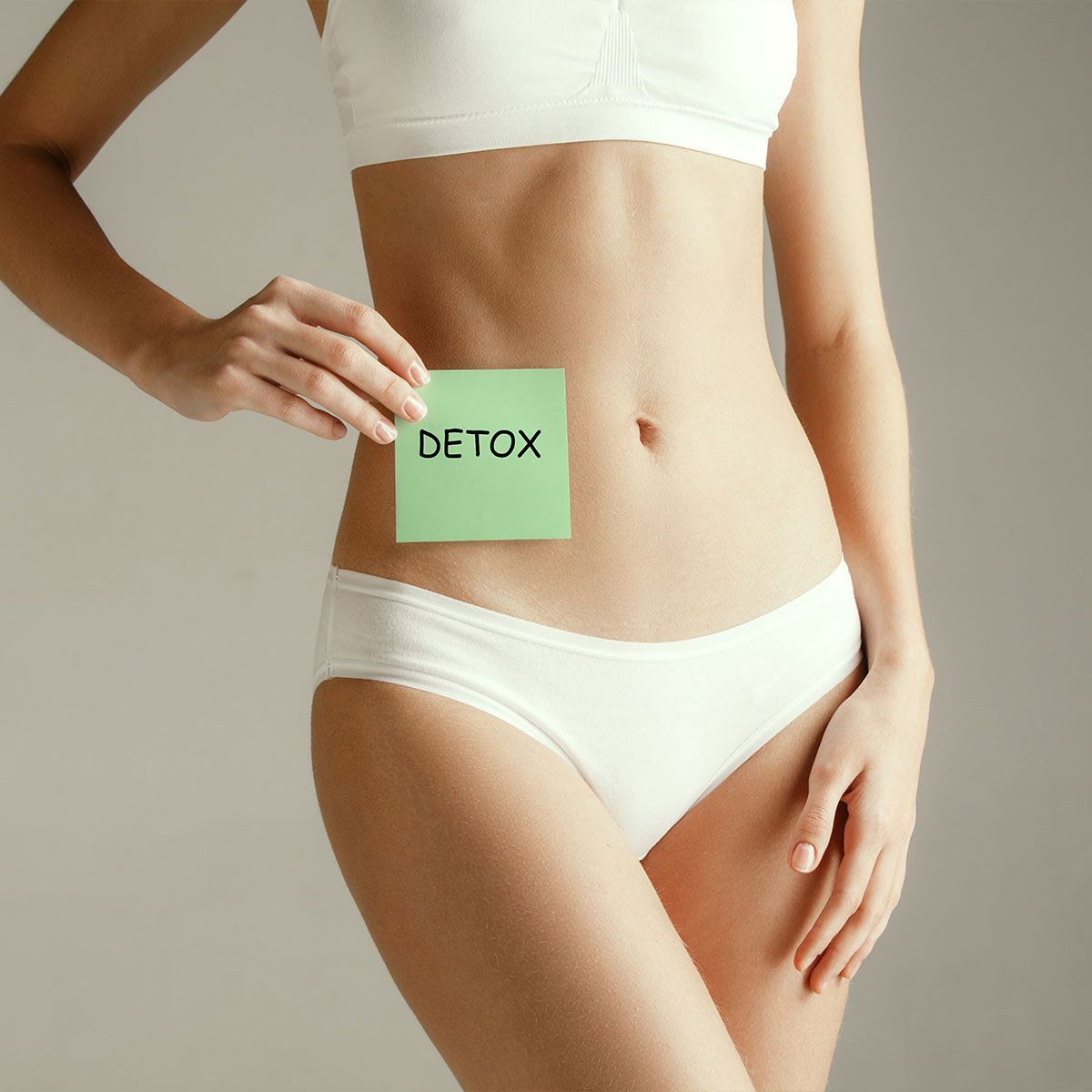 Young adult woman holding card with word DETOX. Woman health. Female model holding card near stomach isolated on gray studio background. Cut out part of body. Medical problem and solution.