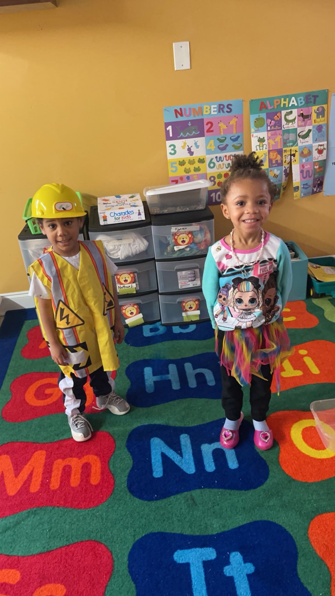 Two young children in costumes stand on a colorful alphabet rug in a classroom. One child is dressed as a construction worker, the other has a Minnie Mouse shirt.