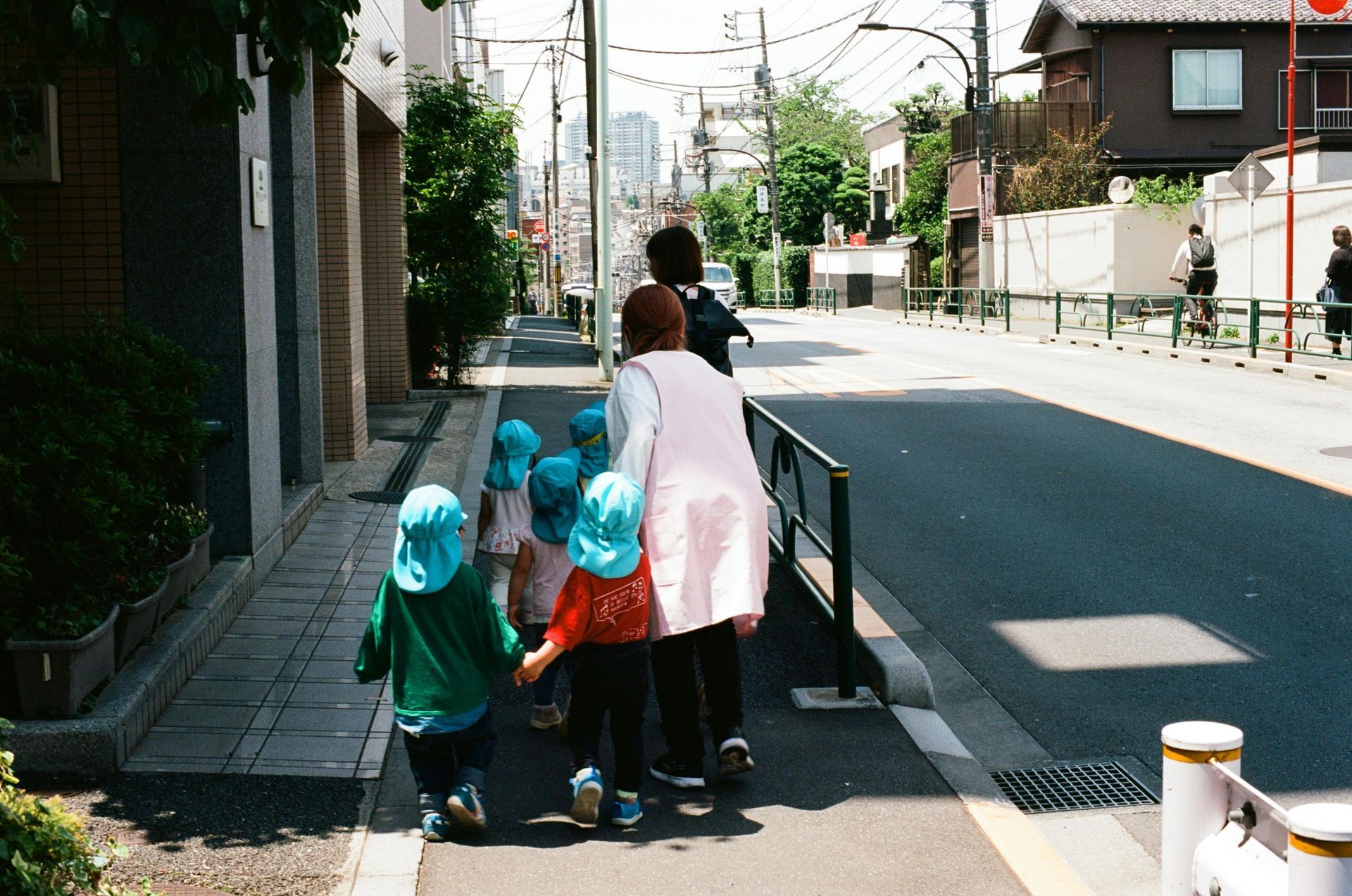 Children in blue hats walk on a sidewalk led by adults in a residential street in Japan.