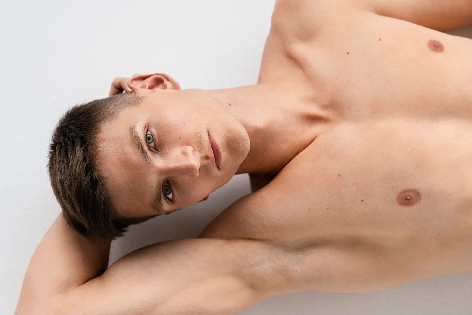 A shirtless young man with dark hair and green eyes looks directly at the viewer while reclining.