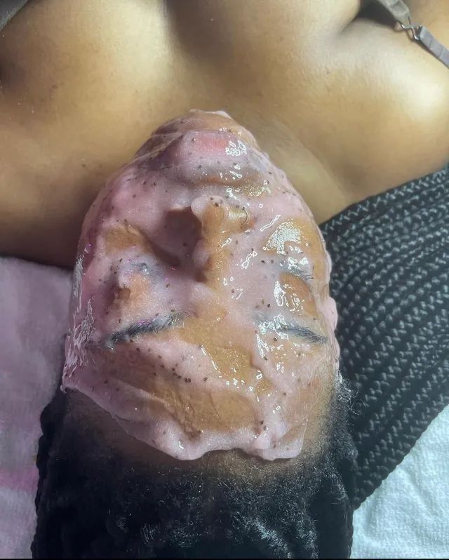 Face covered in pink exfoliating mask. Below are dark eyebrows and part of the person's dark hair, a dark-skinned chest, and dark-patterned clothing.