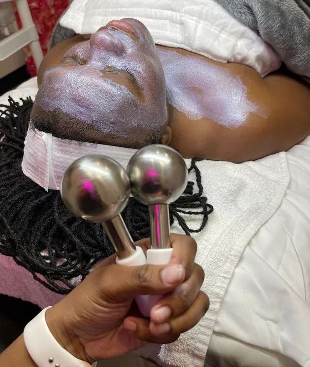 Person receiving a facial treatment with silver electrode tools. A person's hand holds the tools while the face has a glowing cream applied.