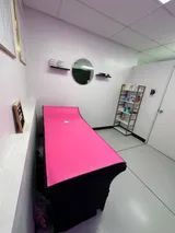 A small spa room with a pink massage table, a round mirror, shelves with products, and a white door.