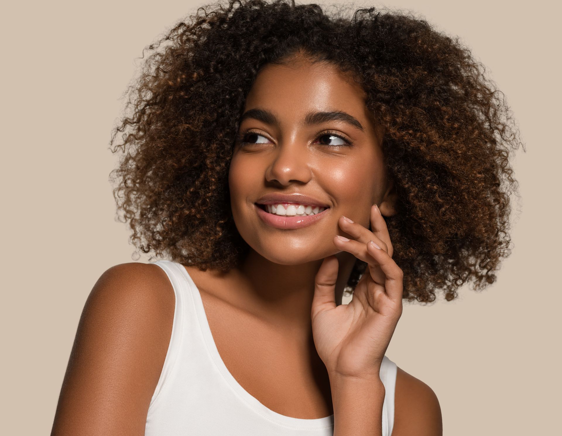 A woman with curly hair is smiling and touching her face.