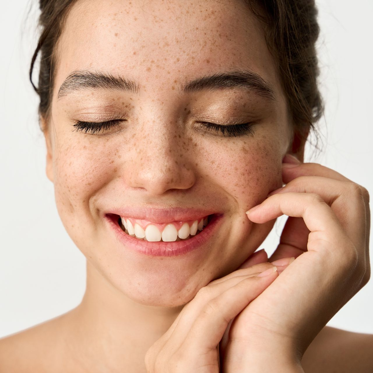 Woman with freckles smiles, eyes closed, hands near face.