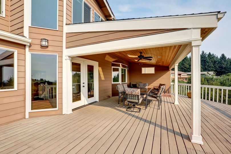 A modern brown house with a covered wooden deck featuring outdoor furniture, a ceiling fan, and views of greenery.