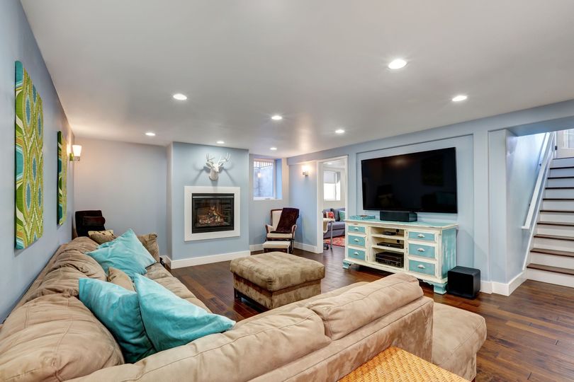 A basement living room with a brown sectional, ottoman, fireplace, large TV on a blue console, and stairs to the right.