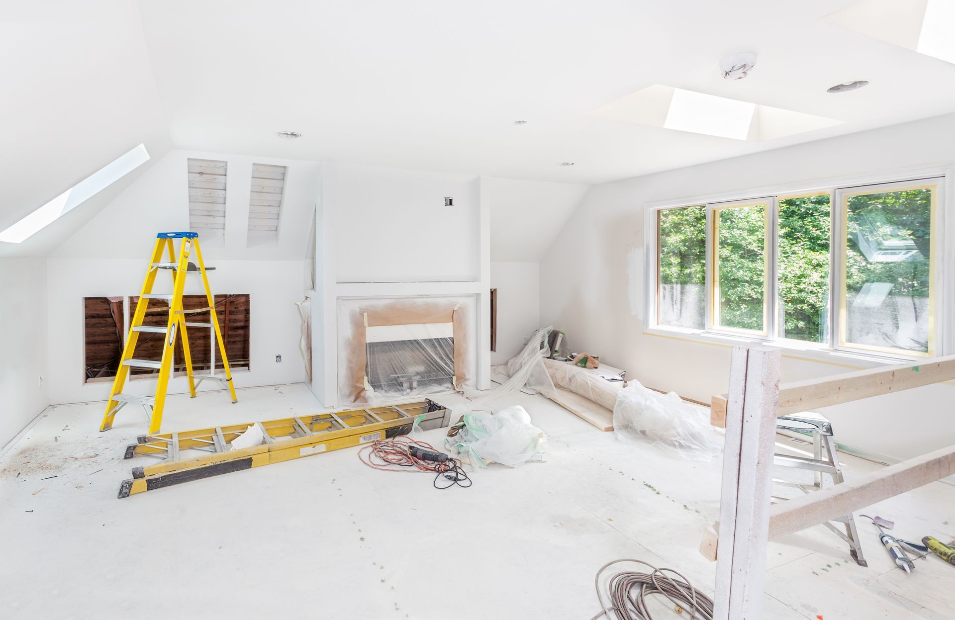 Bright white room under renovation with ladders, tools, plastic-covered furniture, and large windows