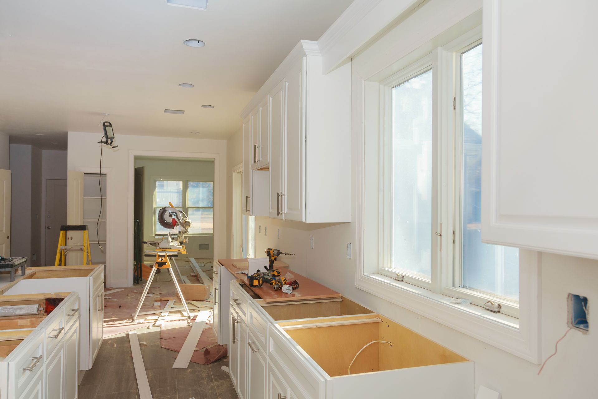 Bright kitchen under renovation with white cabinets, wood floors, and a large window.