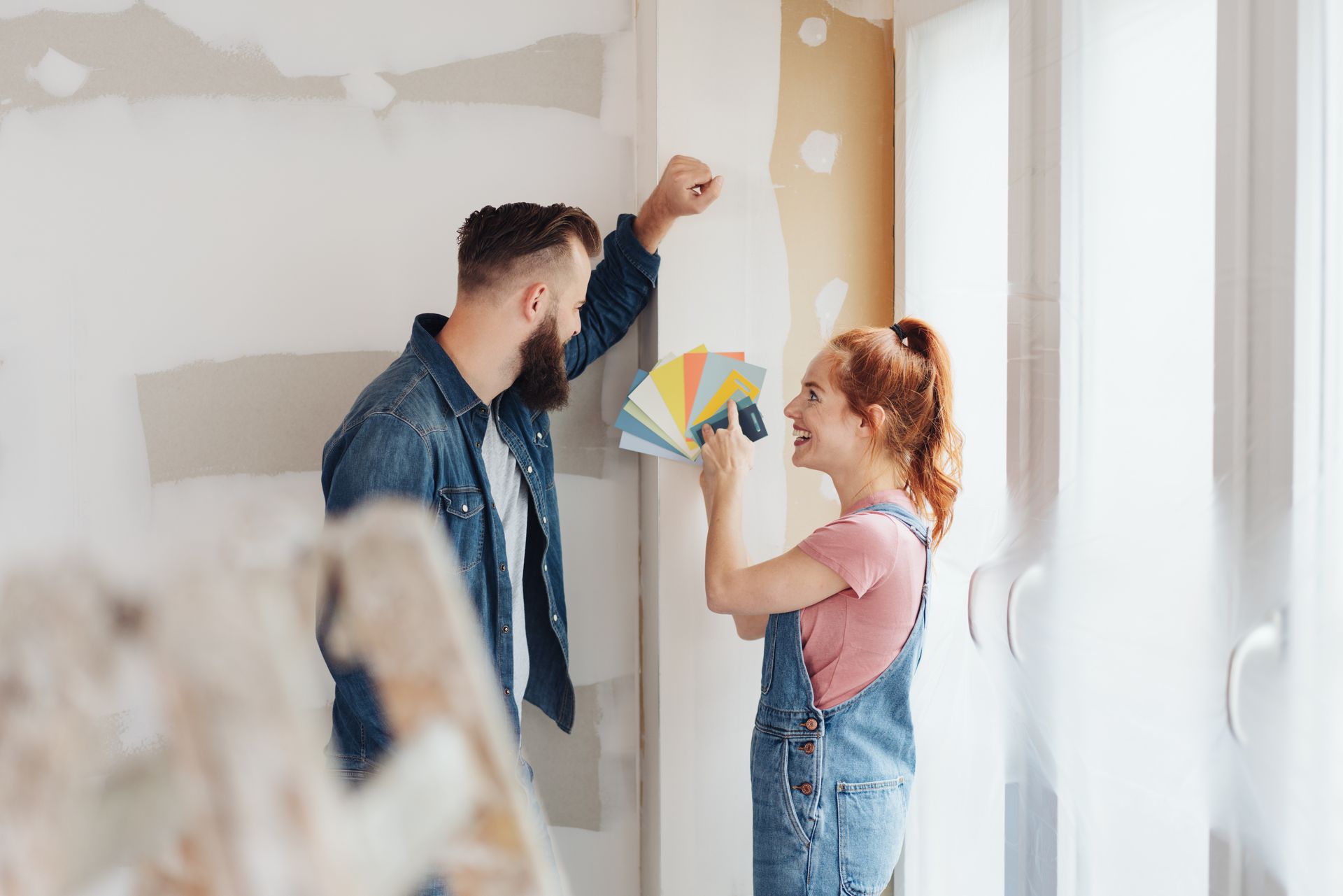 Two people paint a room, with one holding color swatches near a partially finished wall.