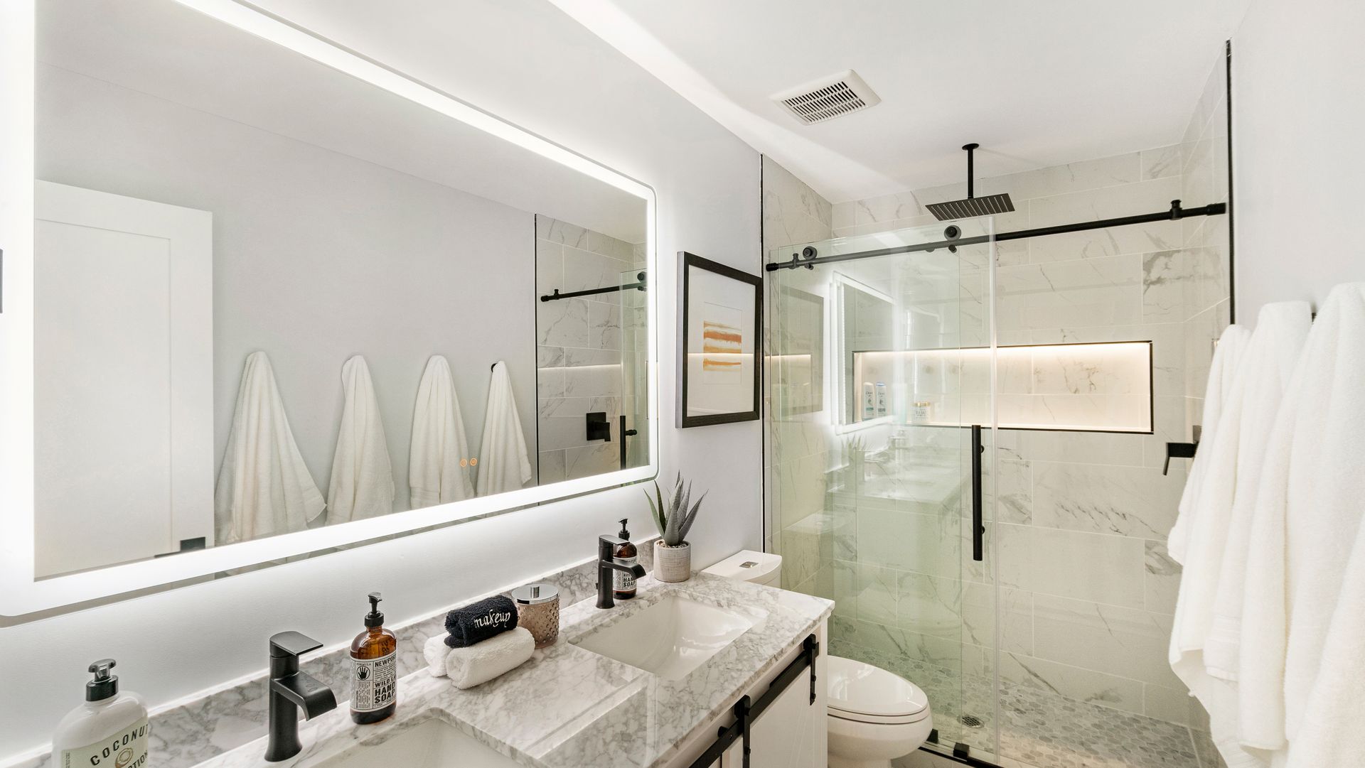 Bright modern bathroom with large mirror, double sink, glass shower, and white towels hanging on the wall.