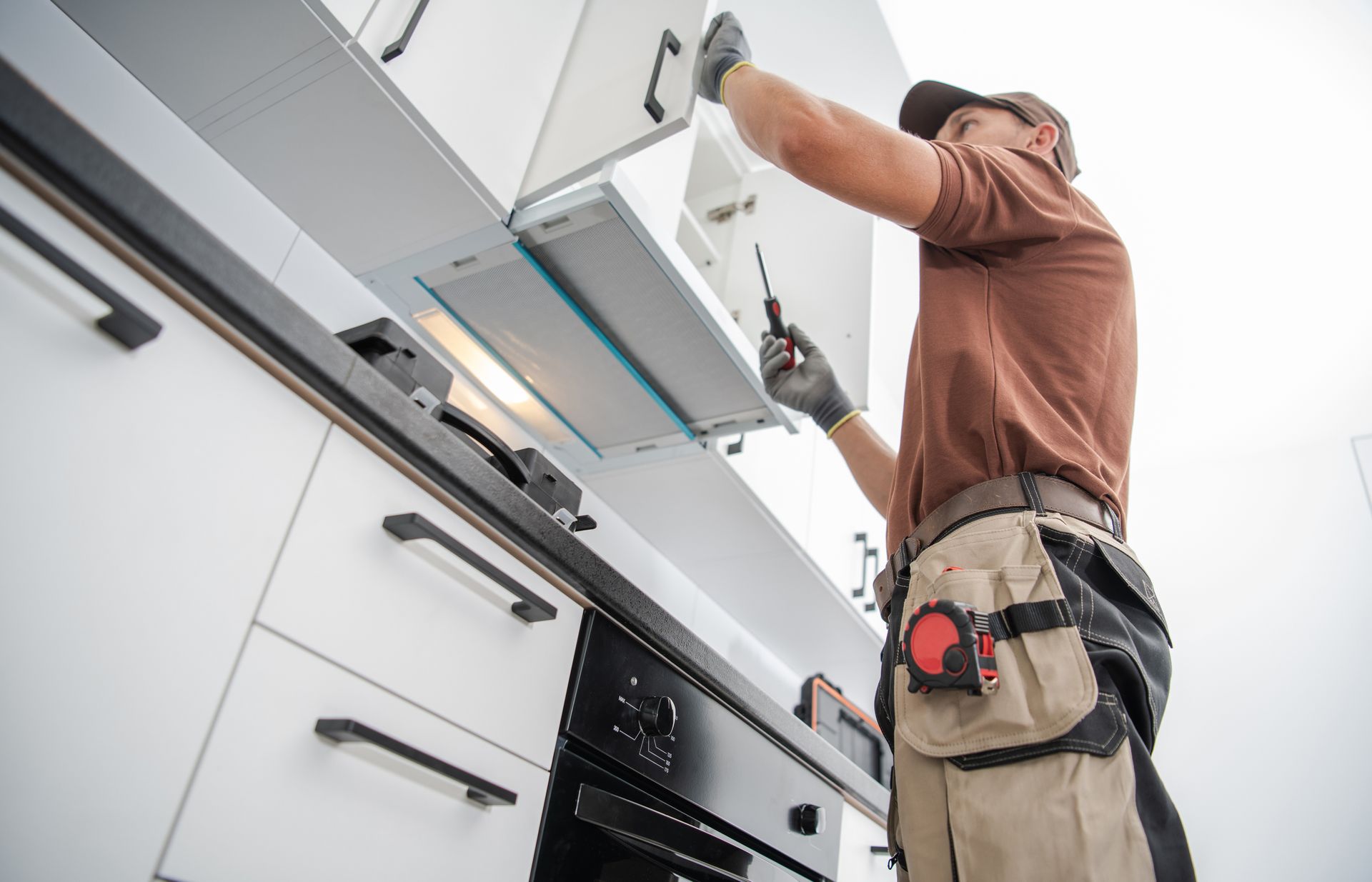 Technician installing a stainless steel kitchen range hood under white cabinets