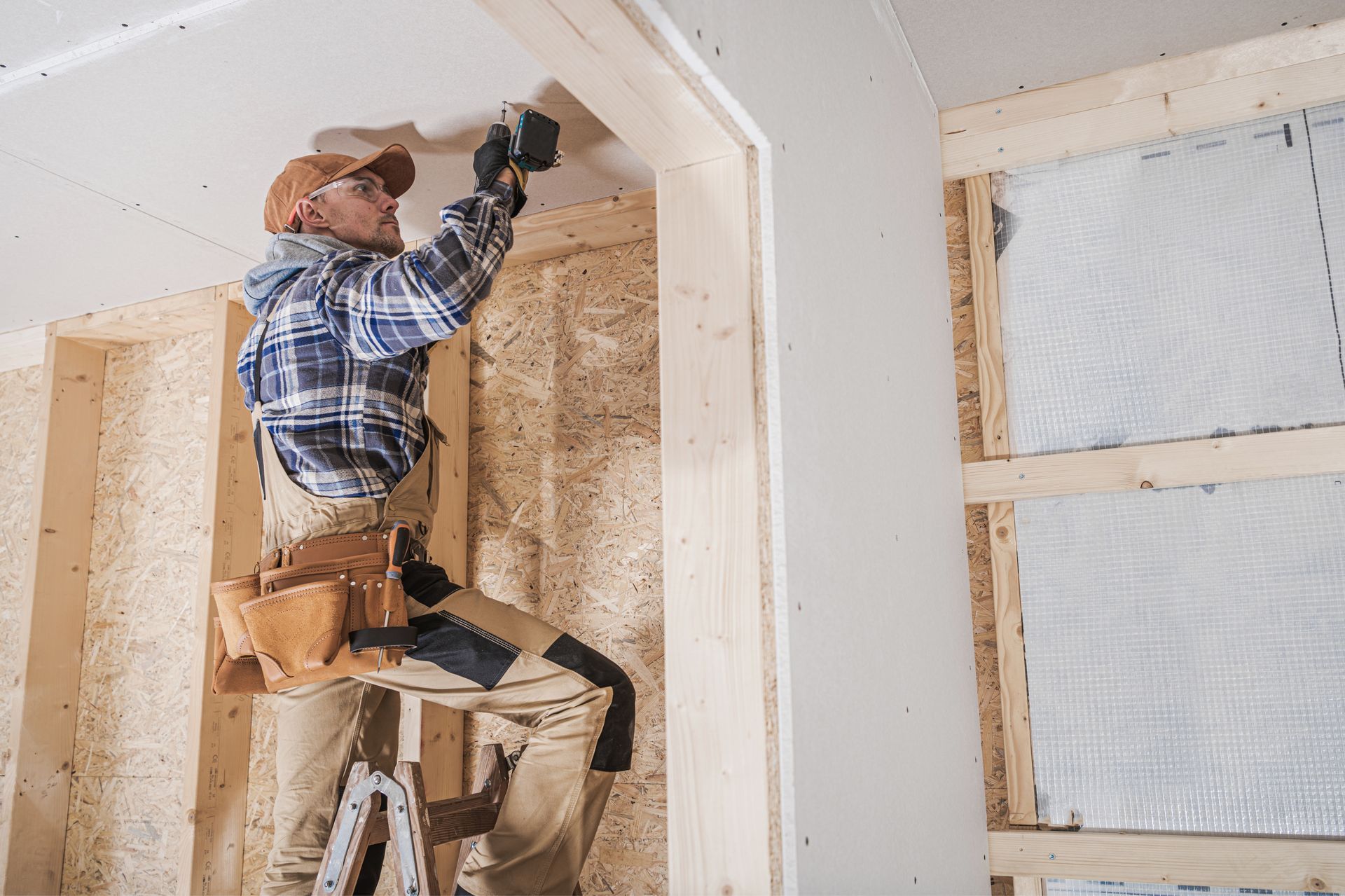 Worker on a ladder installing drywall inside a room with white walls and a window.