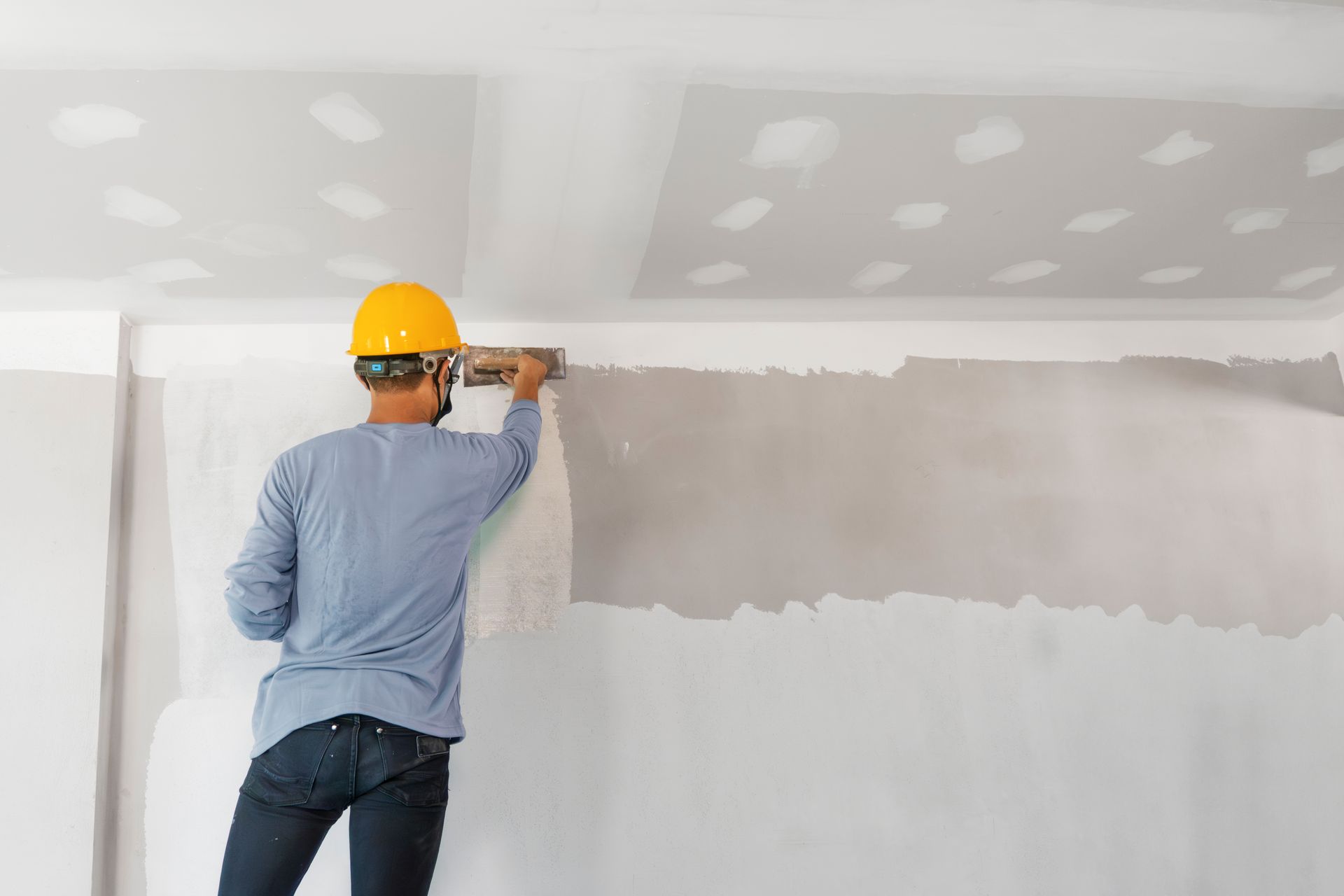 Worker in a yellow hard hat mudding drywall with a trowel in a white room under renovation