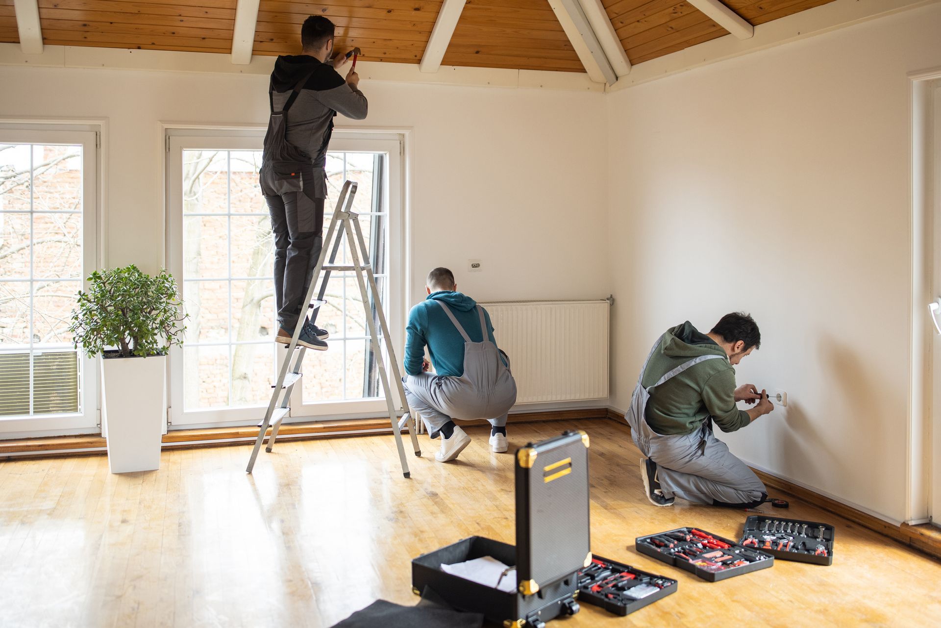 Three workers in workwear perform repairs in a room: one on a ladder, one squatting, and one kneeling by electrical outlets.