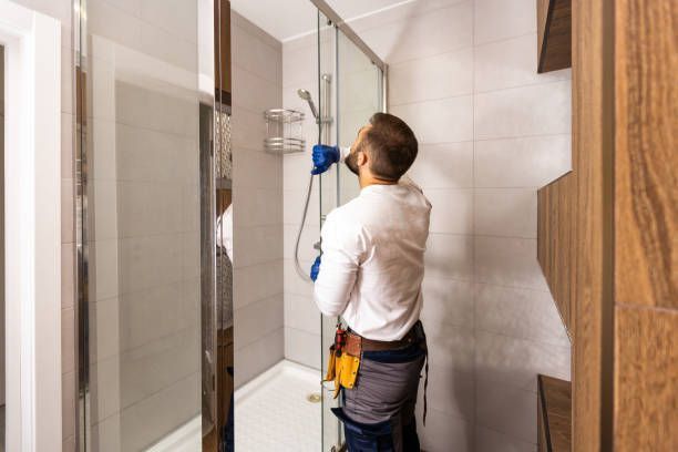 A person in a white long-sleeved shirt and tool belt installs a glass shower door in a tiled bathroom.
