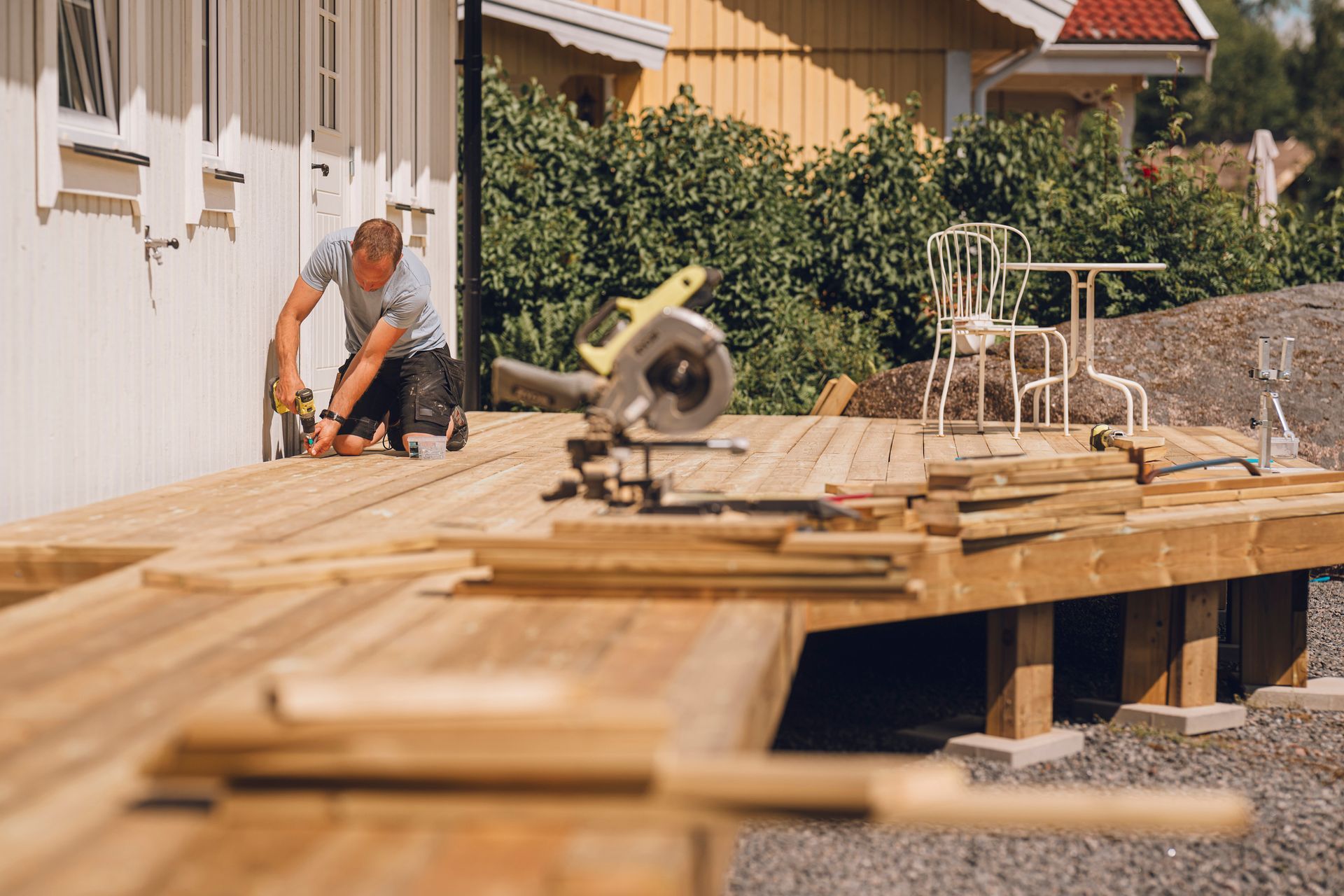 A person works on a wooden deck project next to a house, with a power saw nearby and stacked lumber in the foreground.