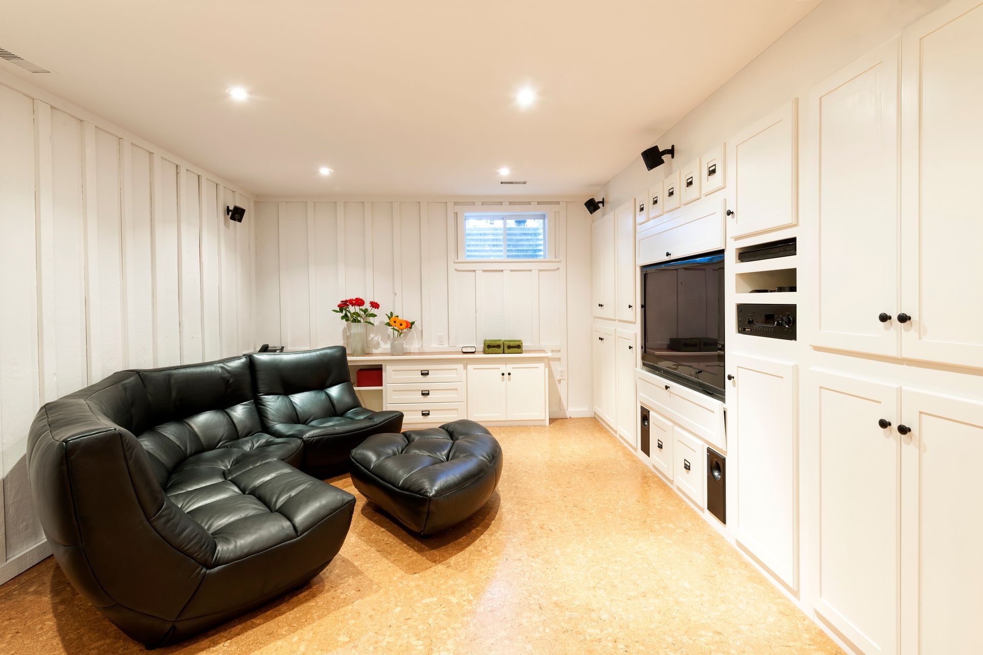 A modern basement den with white paneled walls, a black leather sectional, a television unit, and cork flooring.
