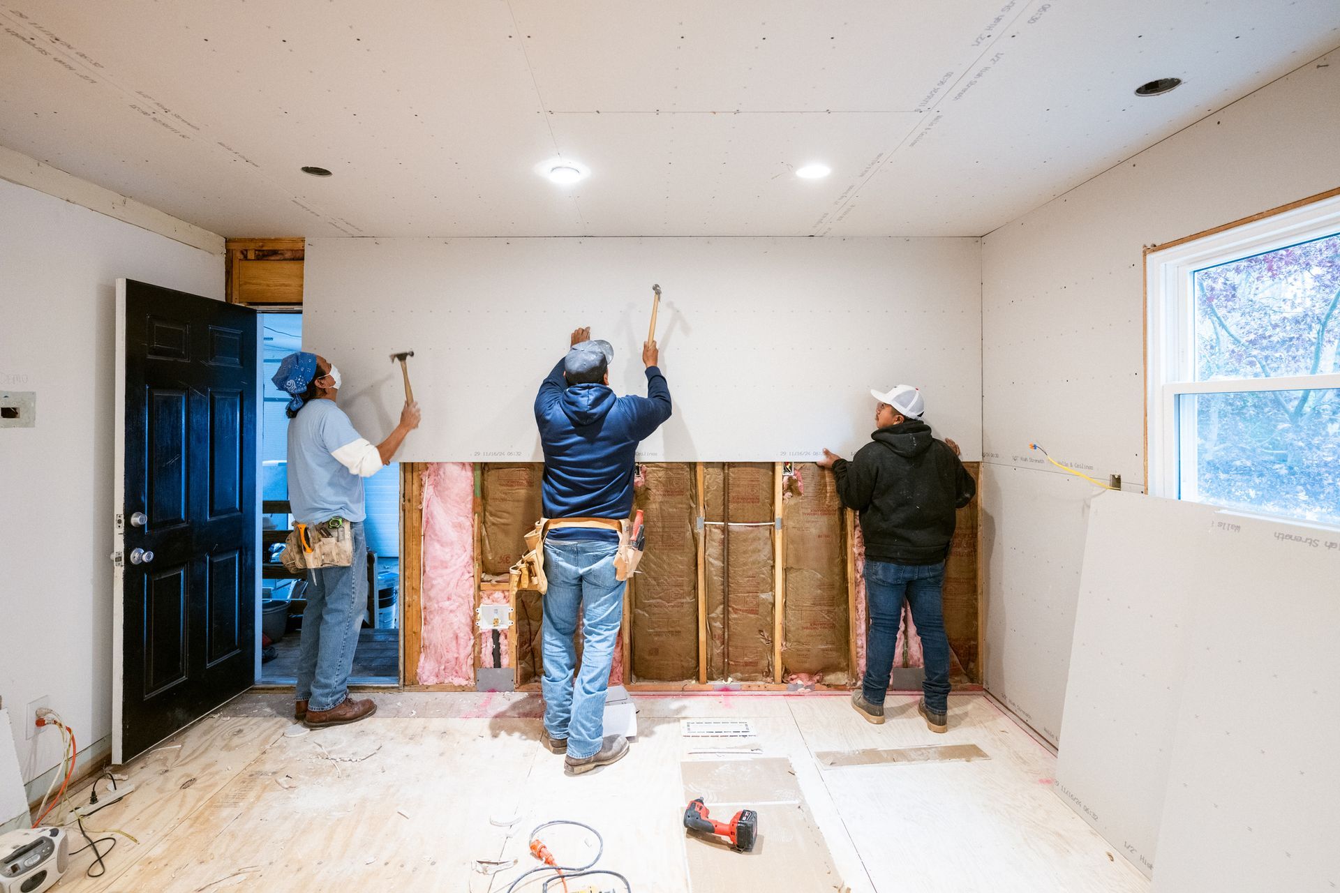 Three workers in casual clothes install drywall panels onto wooden wall studs inside a room under renovation.