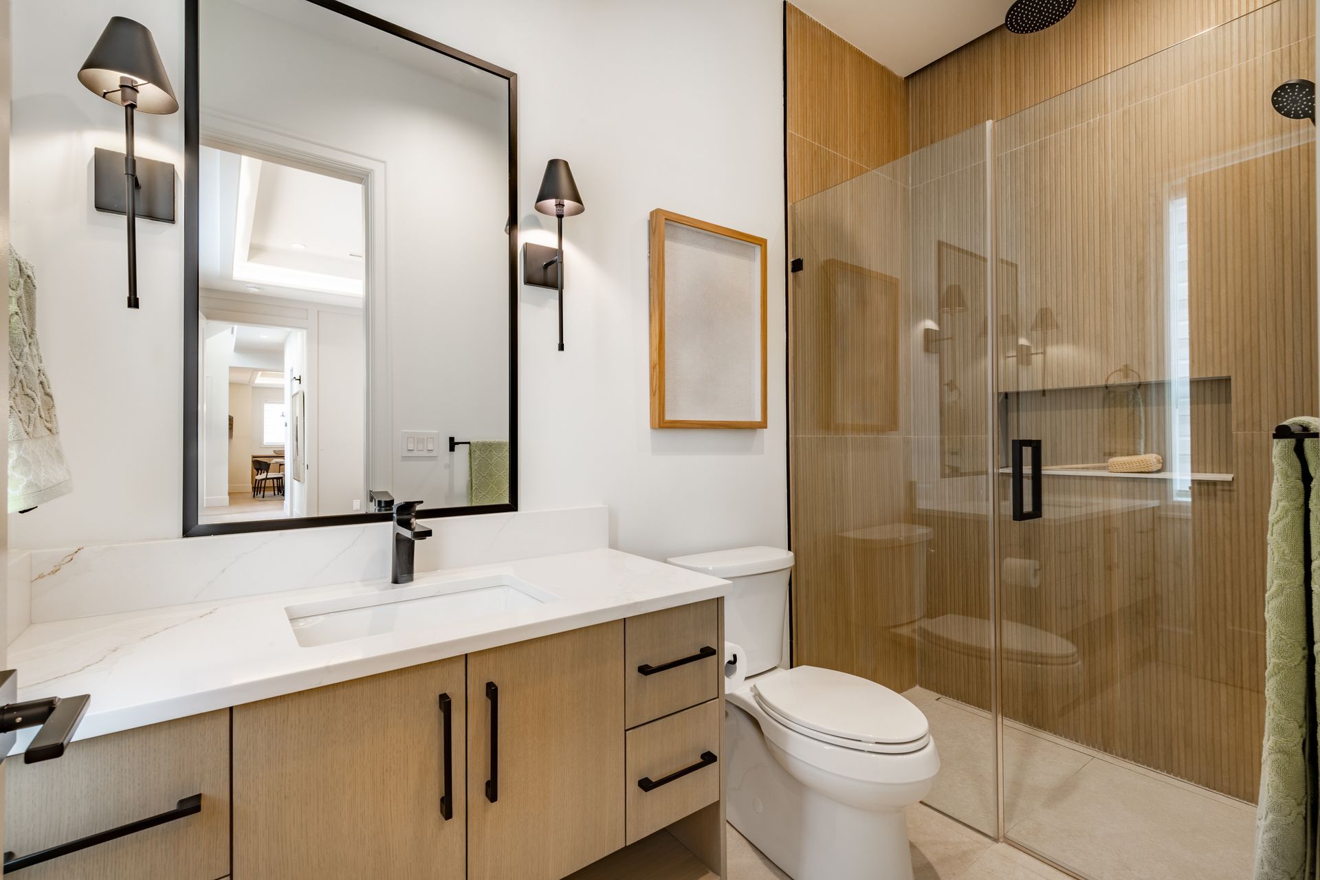 Modern bathroom with a light wood vanity, white countertop, rectangular mirror, wall sconces, and a glass-enclosed shower.