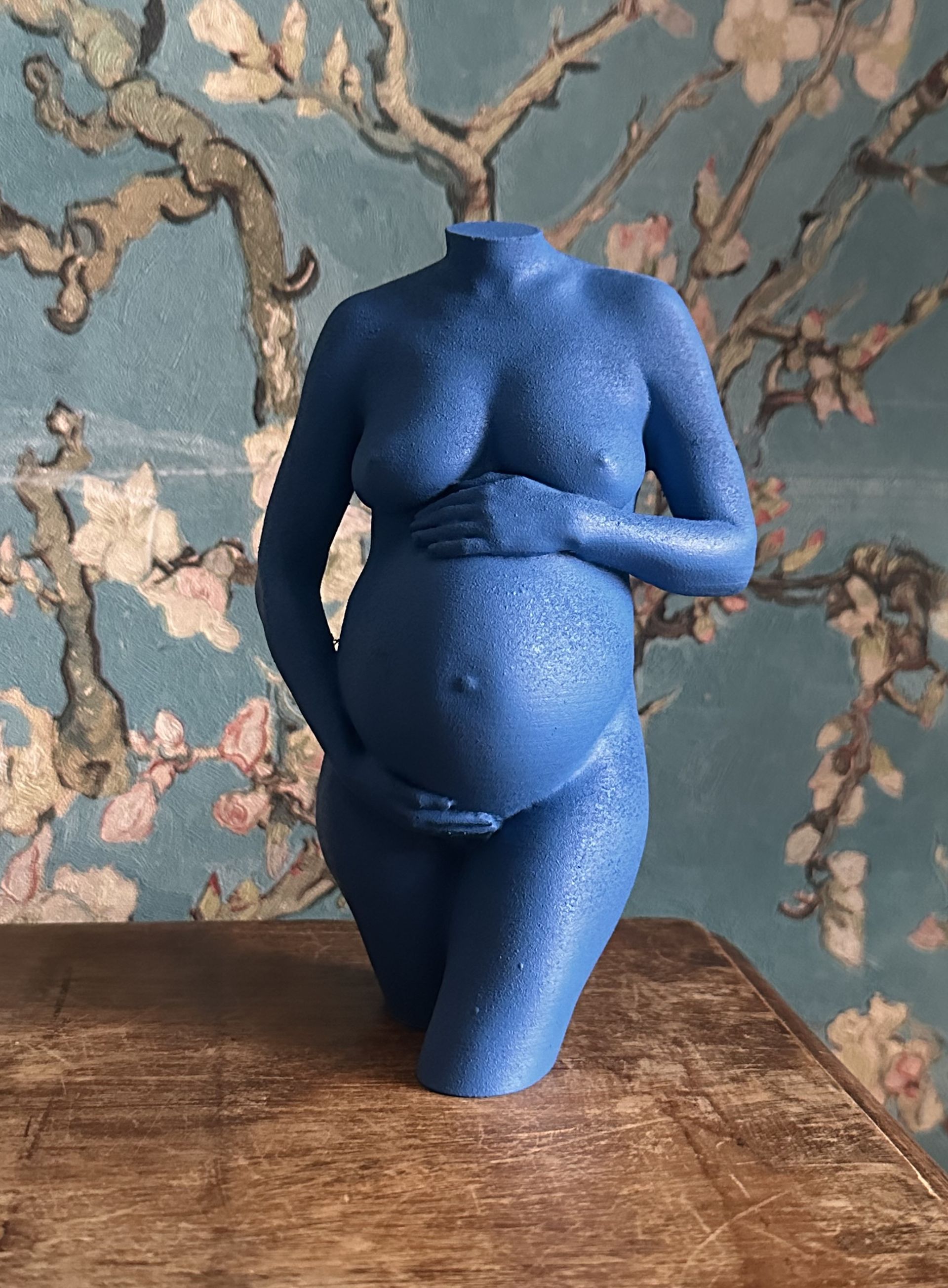 Blauwe torsosculptuur met opgeheven arm op een houten tafel tegen een achtergrond van bloemenbehang.