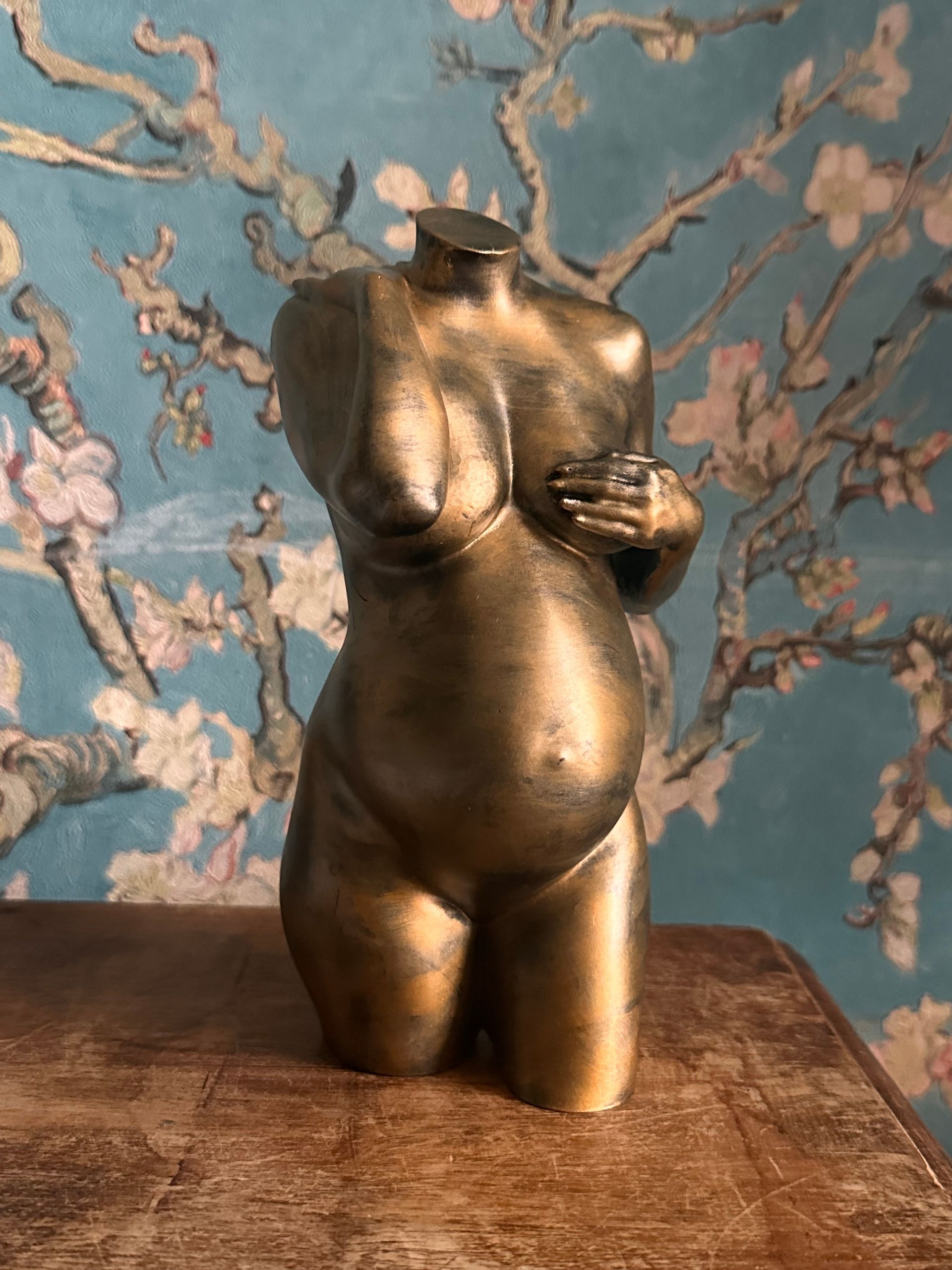 Gouden torso-sculptuur op een houten tafel tegen een blauwe achtergrond met bloemenmotief.