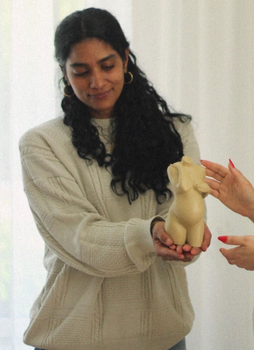 Een vrouw in een beige trui houdt een klein beeldje vast, terwijl een andere hand ernaar reikt.
