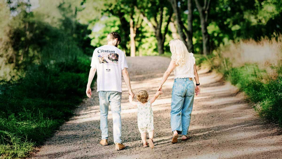 Family walking away on a gravel path, holding hands. A woman and man lead a toddler, trees line the sides of the path.