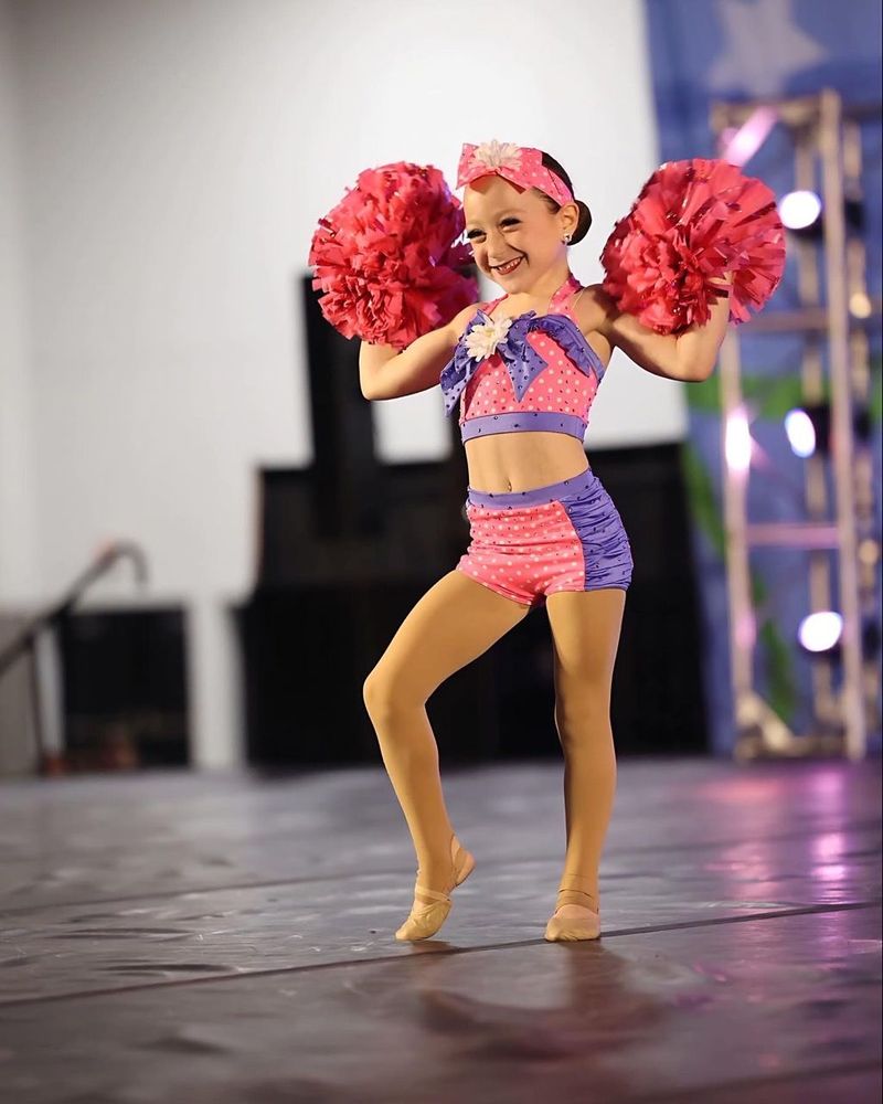 A little girl in a cheerleader outfit is holding pink pom poms