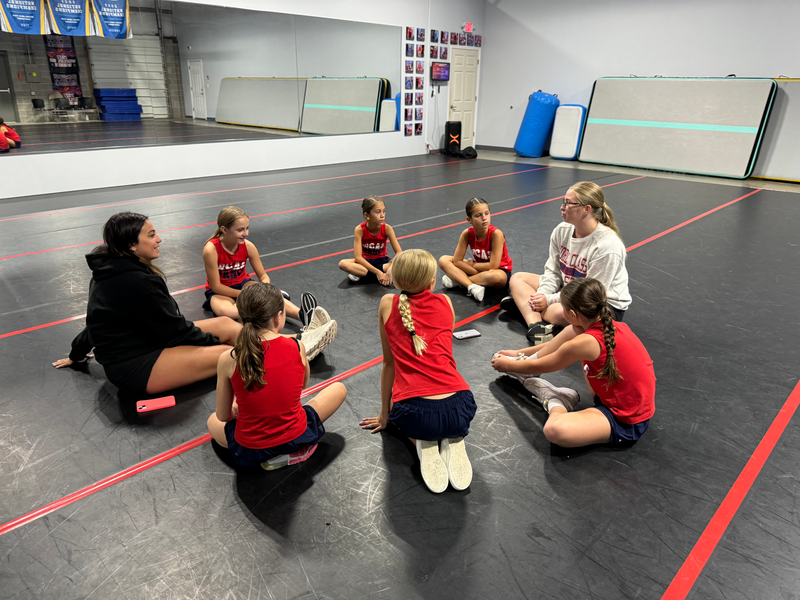 A group of young girls are sitting in a circle on the floor in a gym.