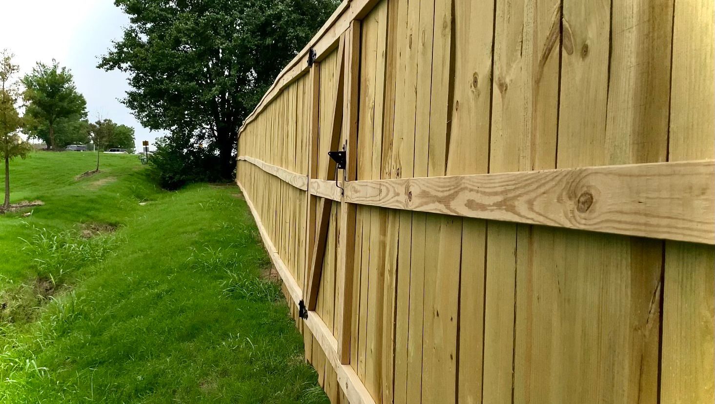 Wooden fence next to a grassy area with trees and a cloudy sky.