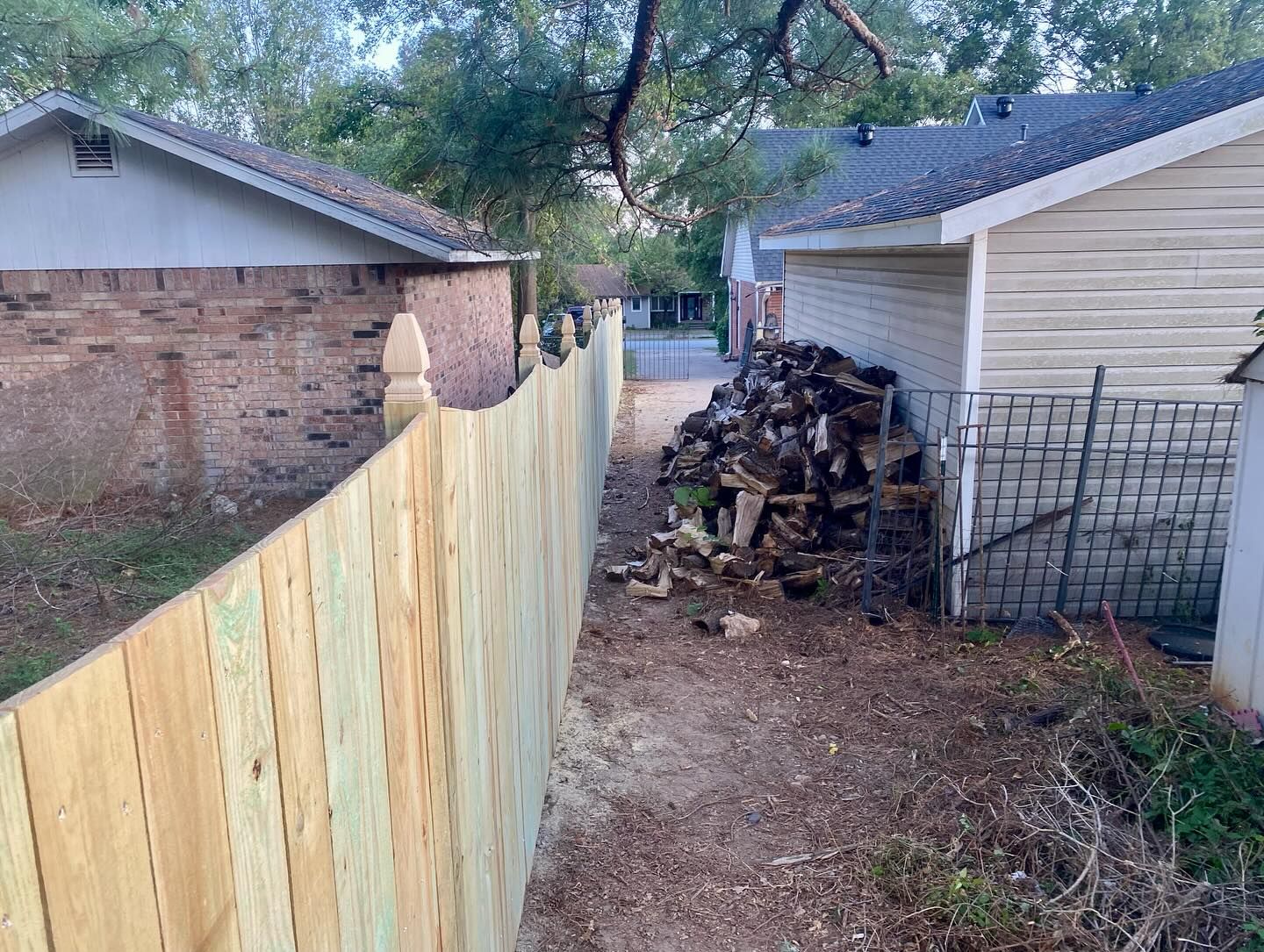 New wooden fence separates two houses, with a pile of firewood visible beside one.