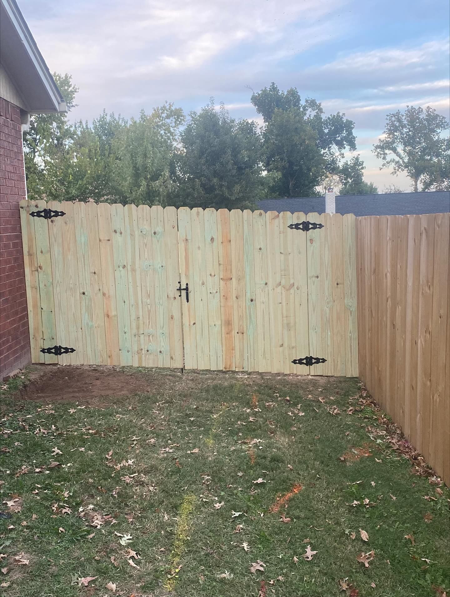 Wooden fence with a gate, attached to a brick wall on the left. Green grass in the foreground.