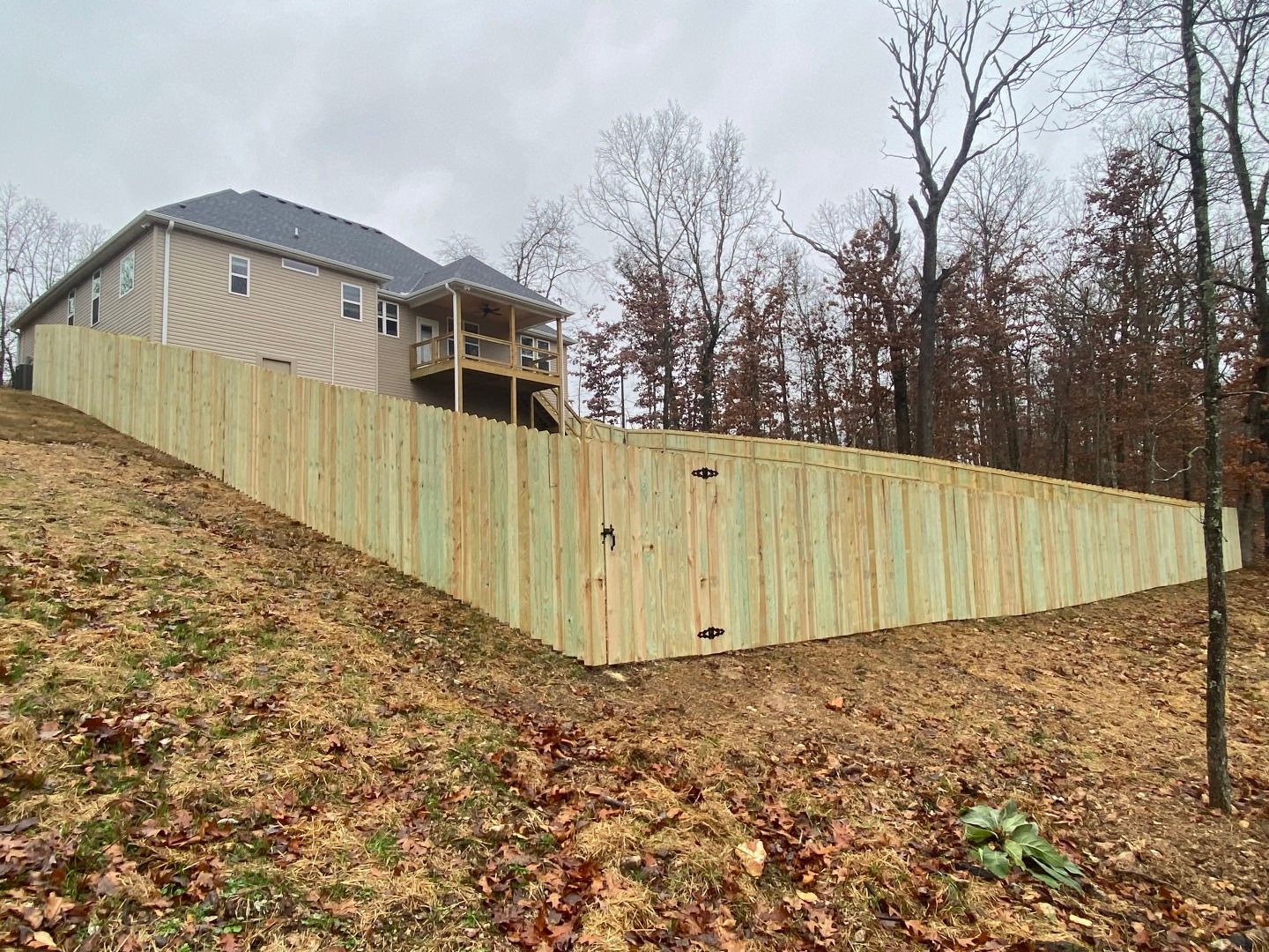 Wooden fence along a sloped yard, next to a two-story house and bare trees.