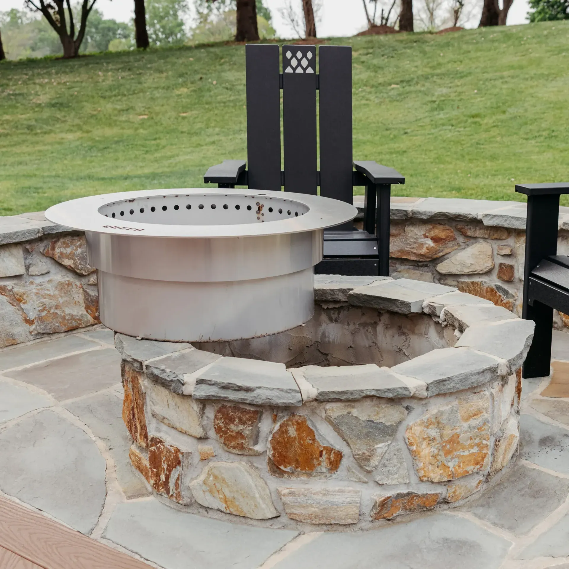 Stone fire pit with metal inserts and a black chair on a patio, with grassy lawn in the background