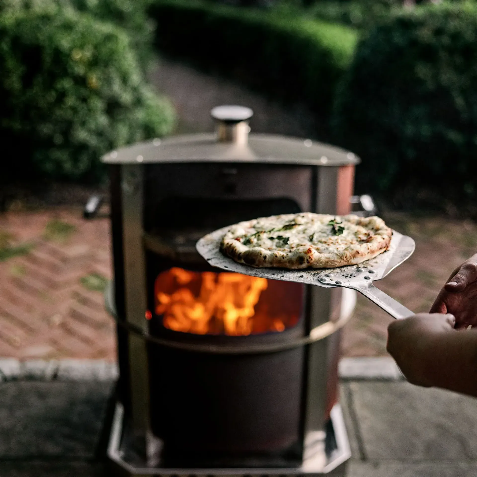 Pizza on a peel being pulled from a wood-fired oven outdoors with flames visible