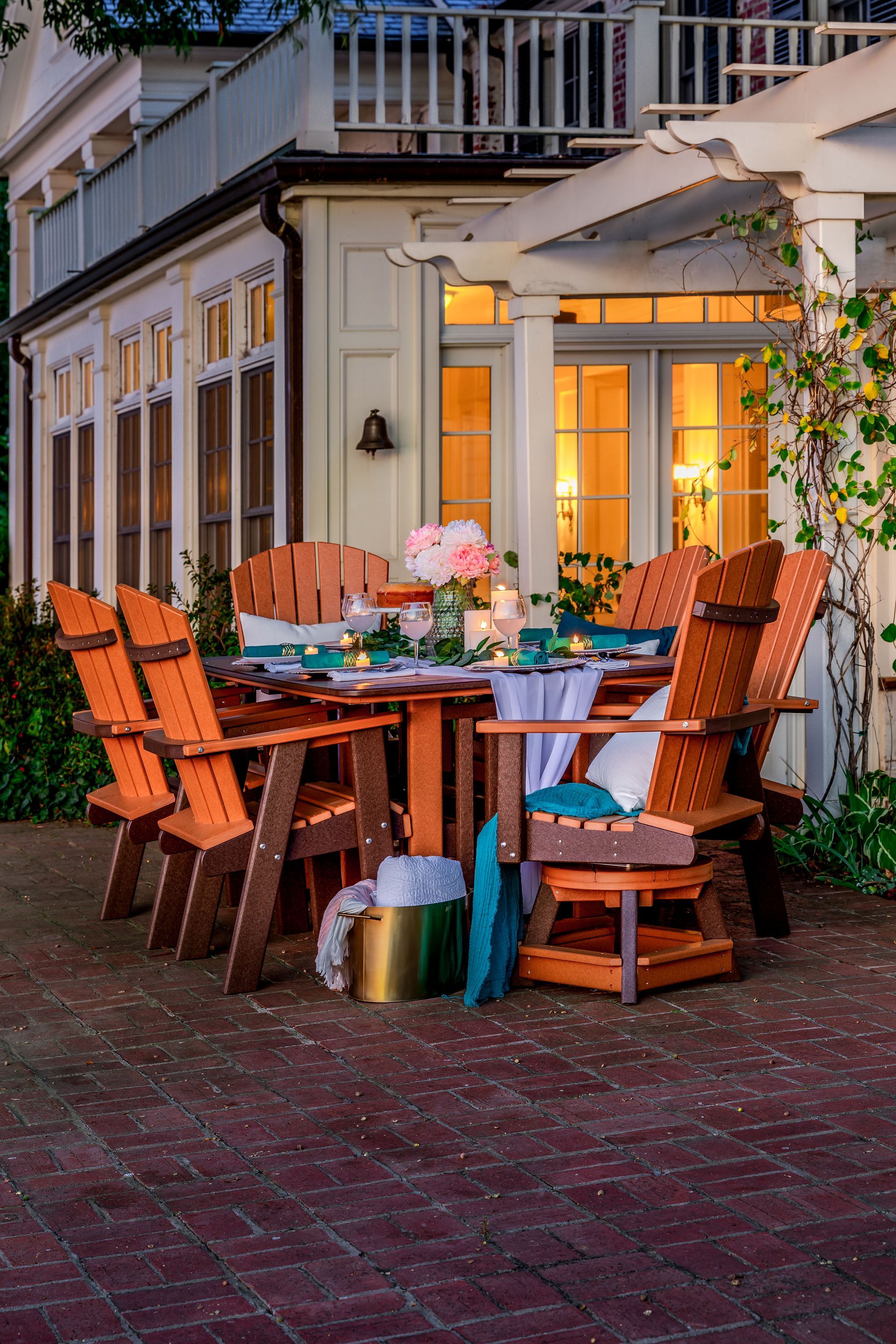 An outdoor patio dining set with wooden chairs and a set table, arranged on brick pavers beside a house at dusk.