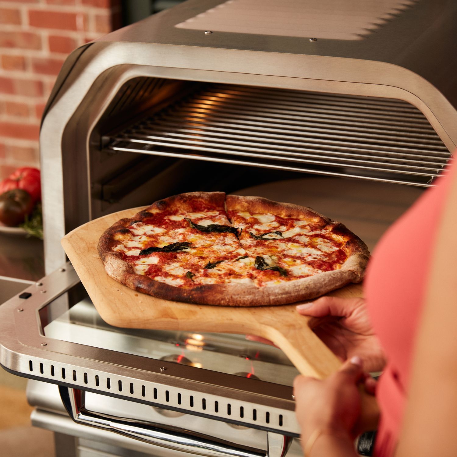 A person holding a pizza peel with a fresh pizza in front of a stainless steel outdoor pizza oven.