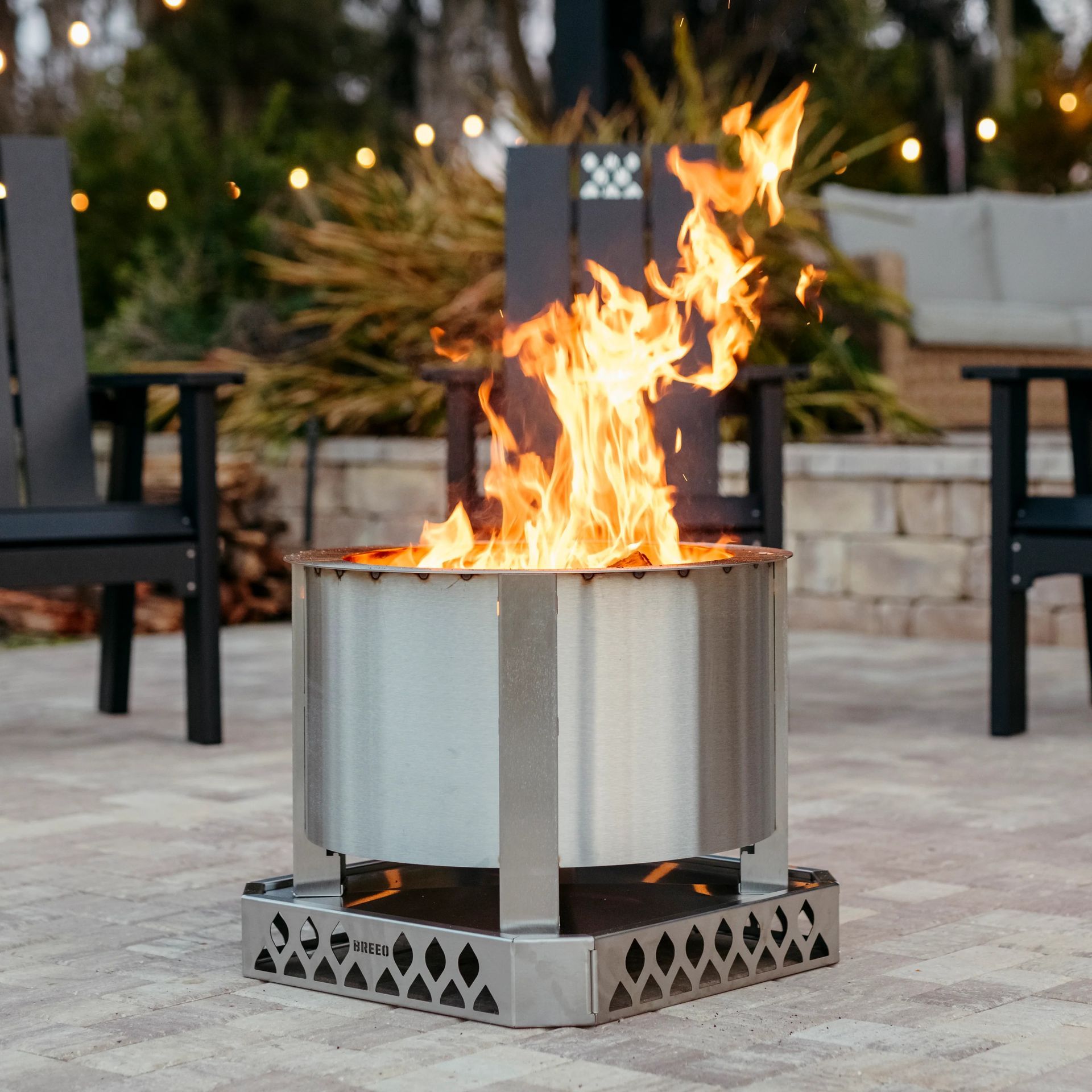 A cylindrical metal fire pit with bright flames burning on a stone patio surrounded by outdoor chairs.
