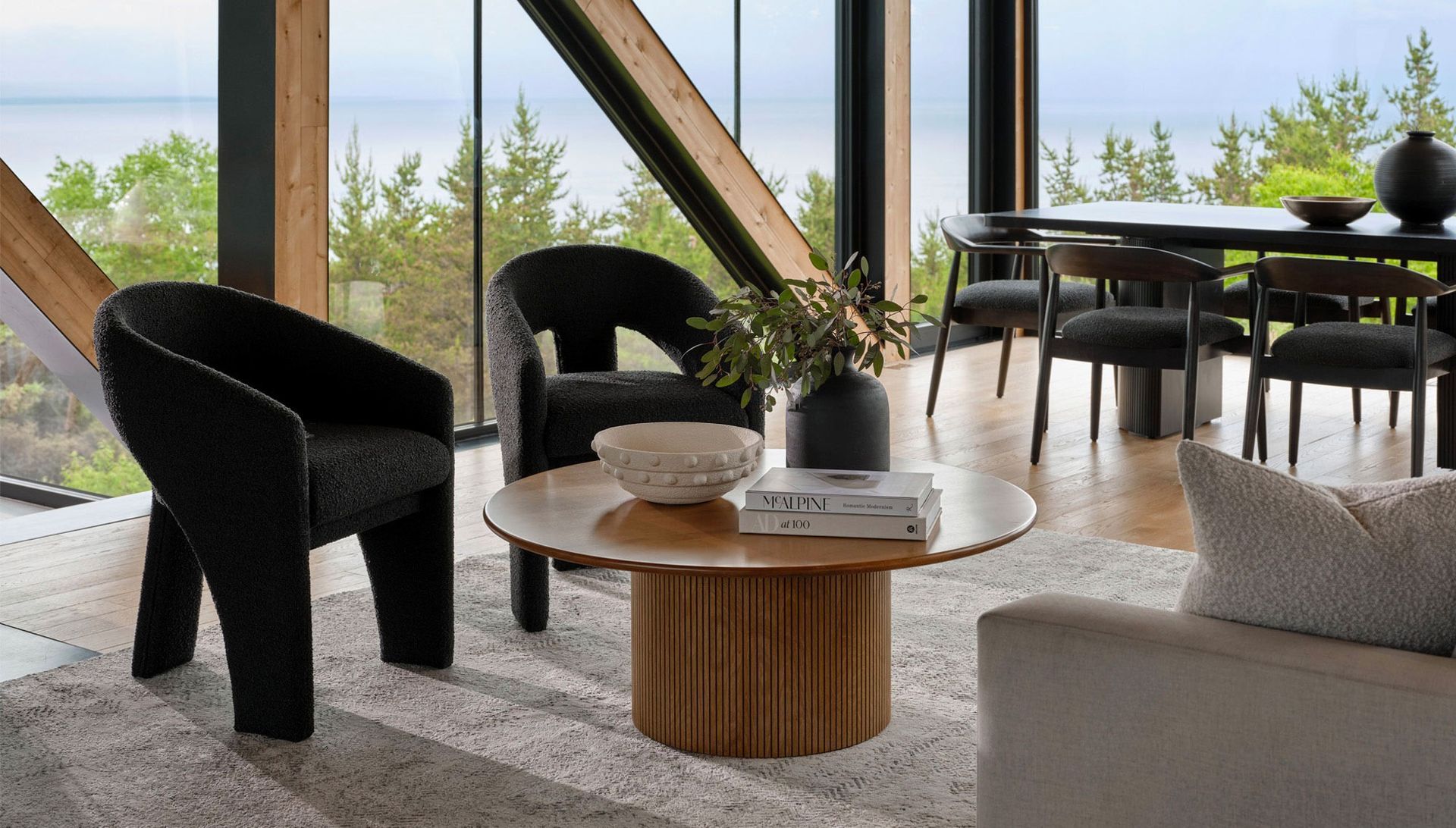Modern living room with a round wooden coffee table, black textured chairs, and a dining area overlooking the outdoors.