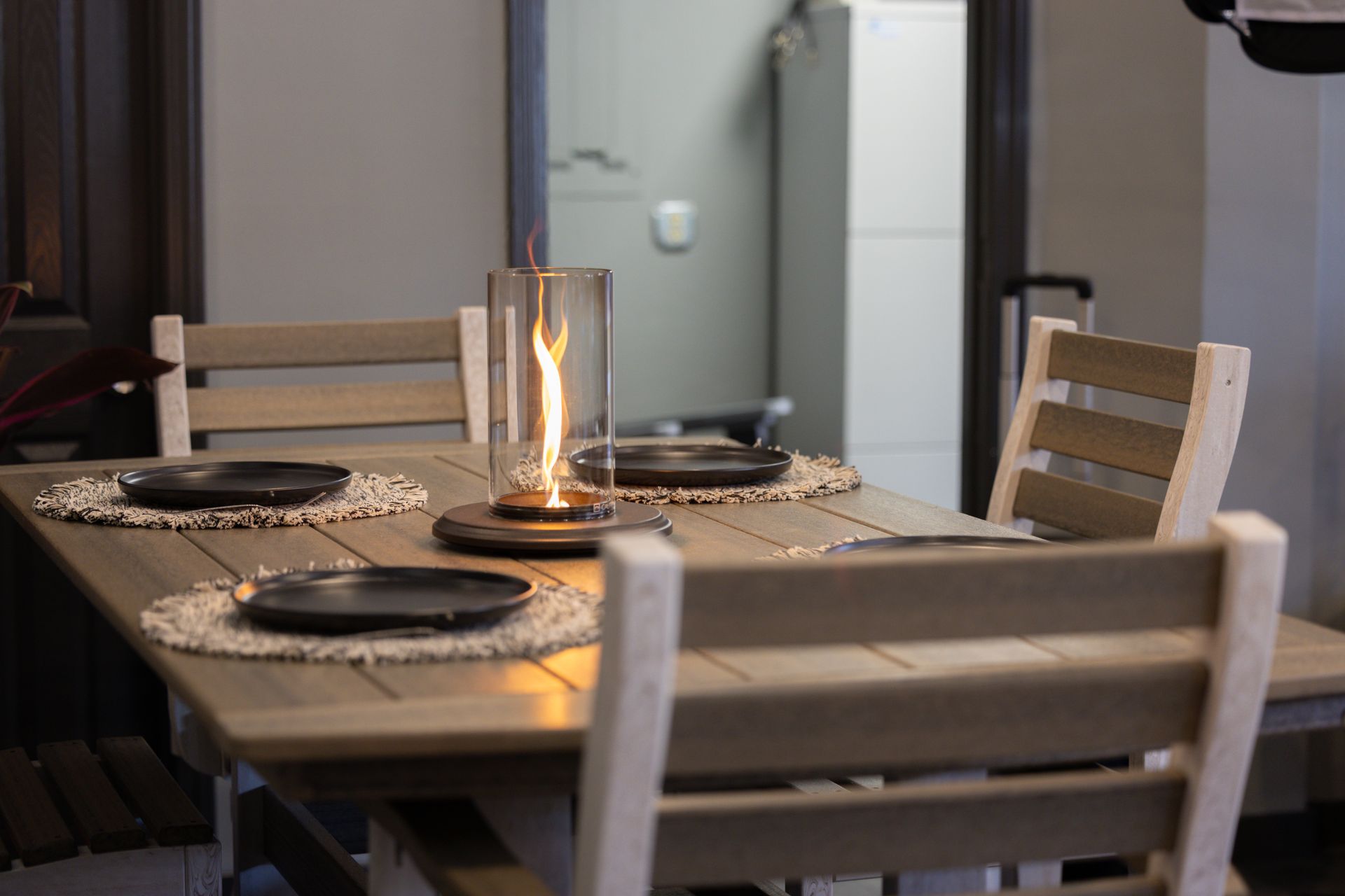 A light wood dining table set for four with woven placemats, dark plates, and a glass-enclosed tabletop fire pit.