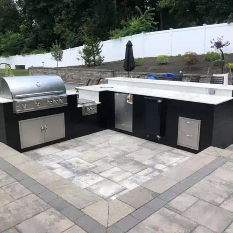 L-shaped outdoor kitchen with a stainless steel grill, counter, cabinets, and stone pavers on a backyard patio.