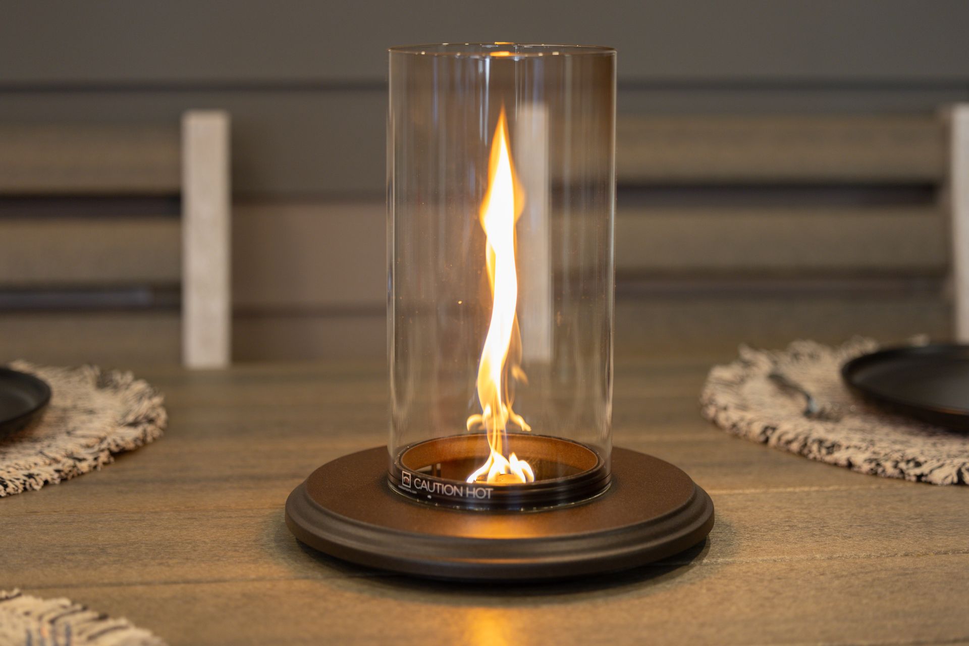 A tabletop fire pit with a single, flickering flame inside a clear cylindrical glass shield, set on a dark wood table.