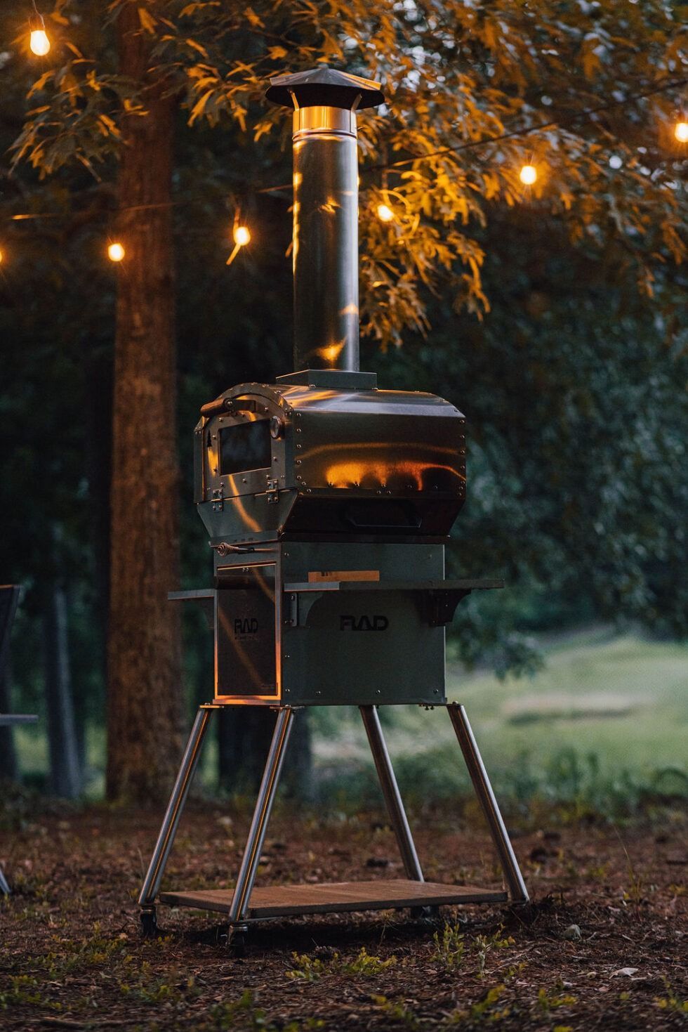 Black outdoor wood stove on tripod in a wooded yard with warm string lights at dusk